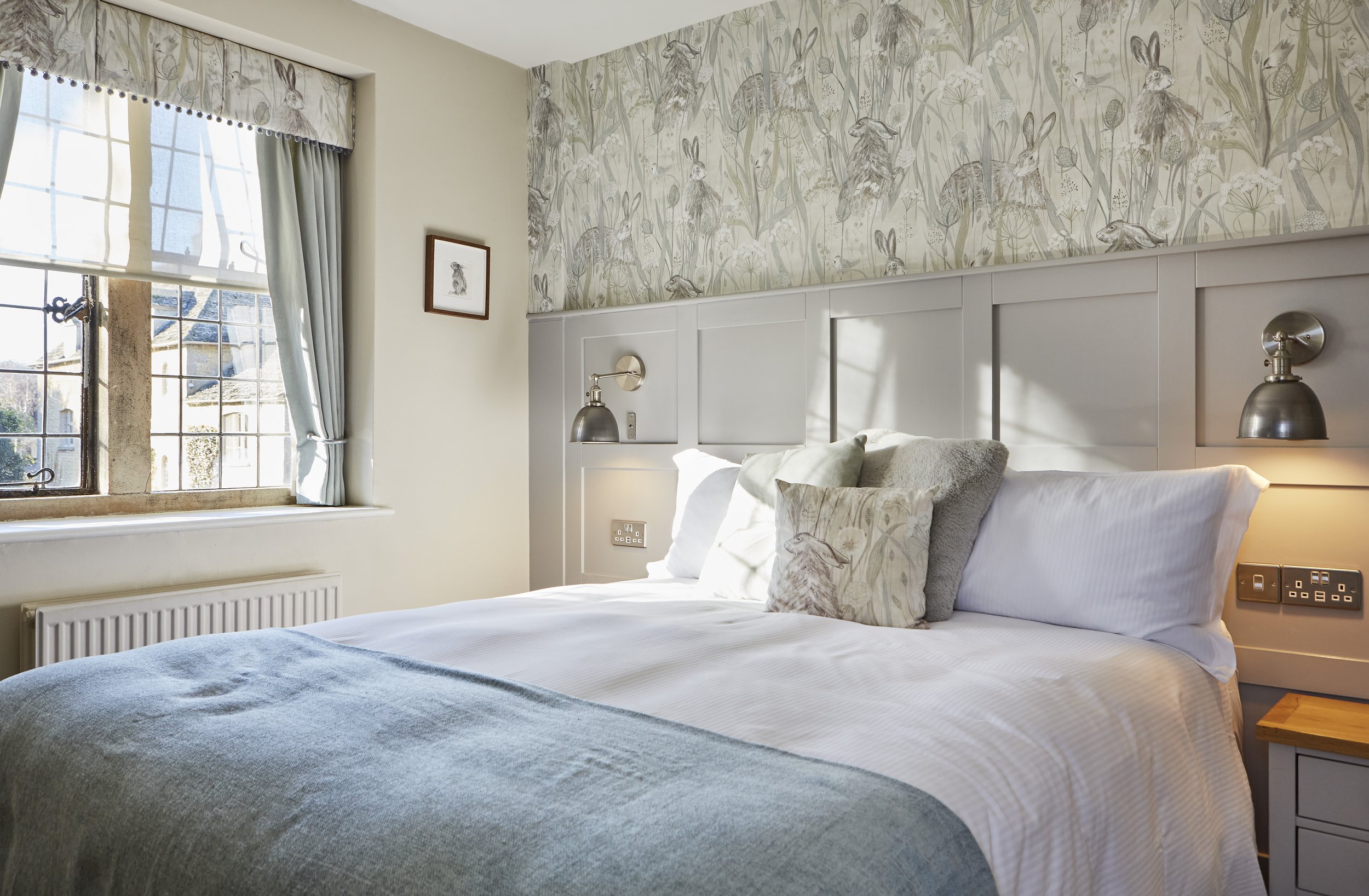 Cozy bedroom with large bed, gray and white bedding, pillow with rabbit design, floral wallpaper with rabbits, window with curtains, framed art, and bedside lamps.