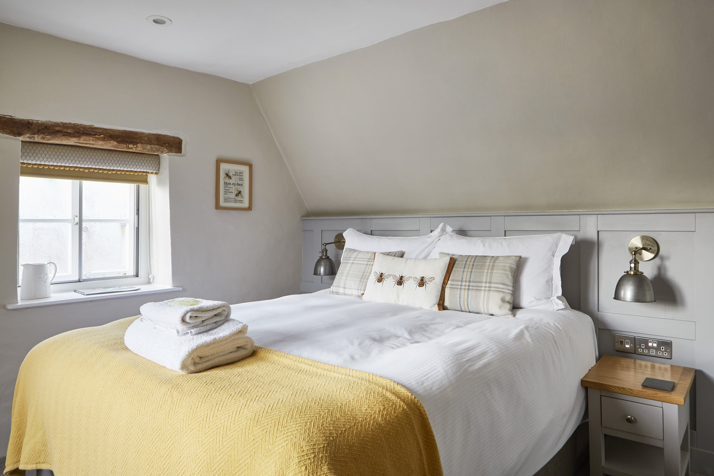 Cozy bedroom with a double bed, adorned with white sheets, patterned pillows, and a yellow throw blanket. Fresh towels are neatly stacked on the bed. A window with a wooden beam above, a small bedside table with a lamp, and a framed picture add charm
