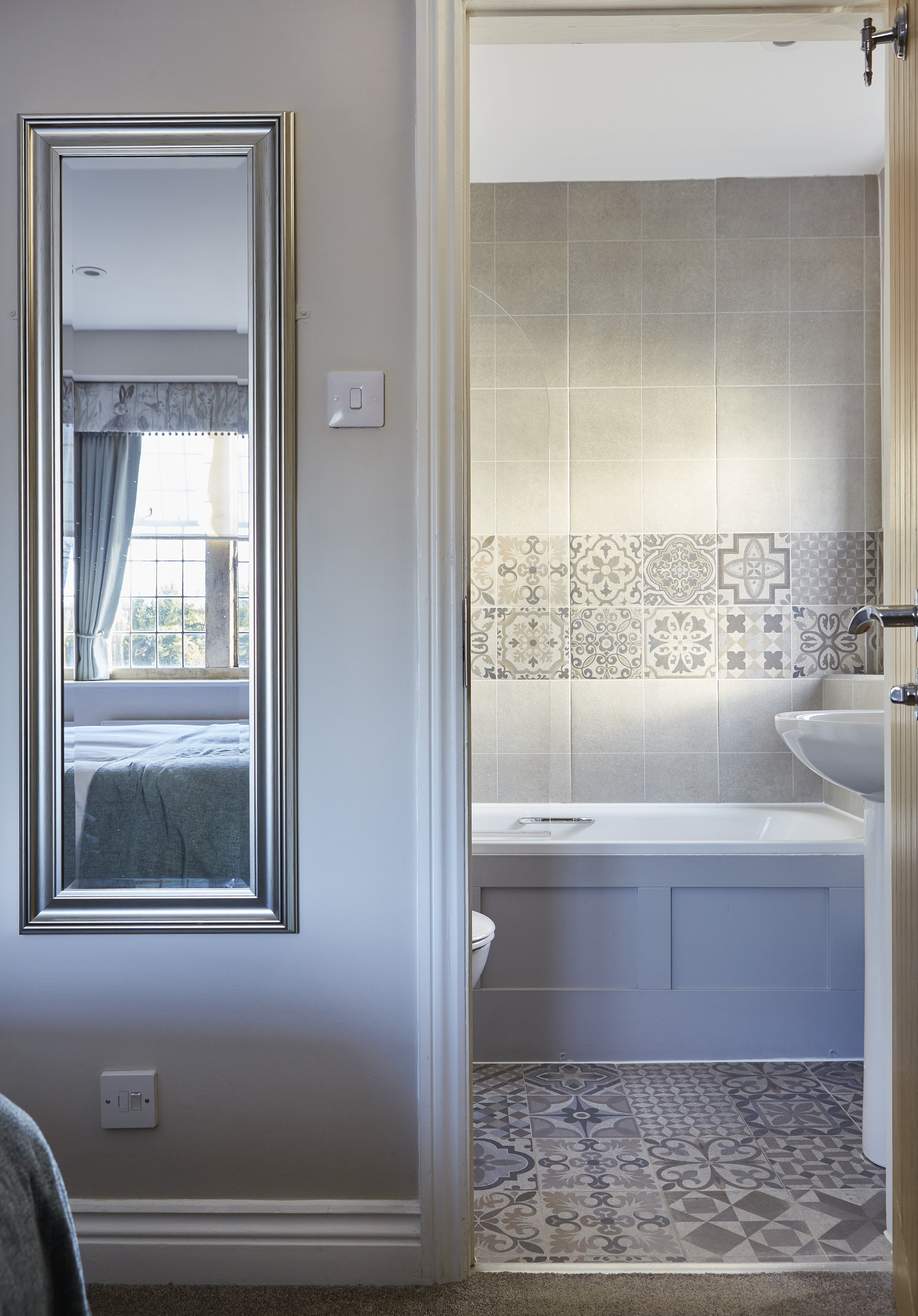Bathroom with patterned tiles, sink, and bathtub; mirror reflecting a bedroom with bed and window.