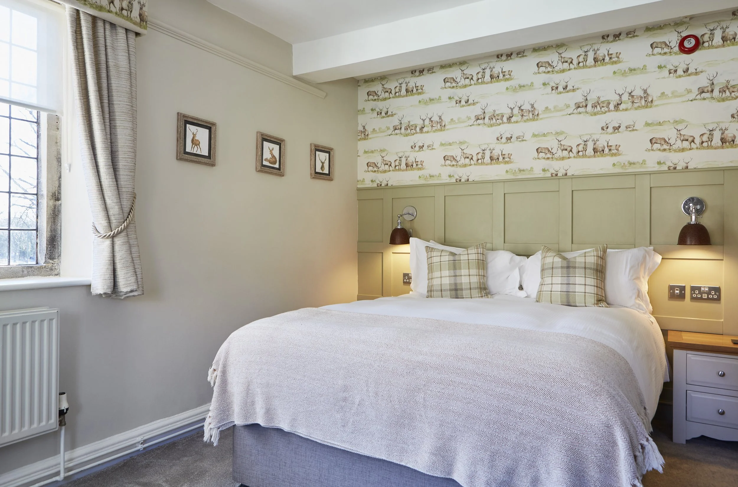 Cozy hotel room with a double bed, plaid pillows, and deer-themed wallpaper. Three small framed pictures hang on a light-colored wall next to a window with curtains.
