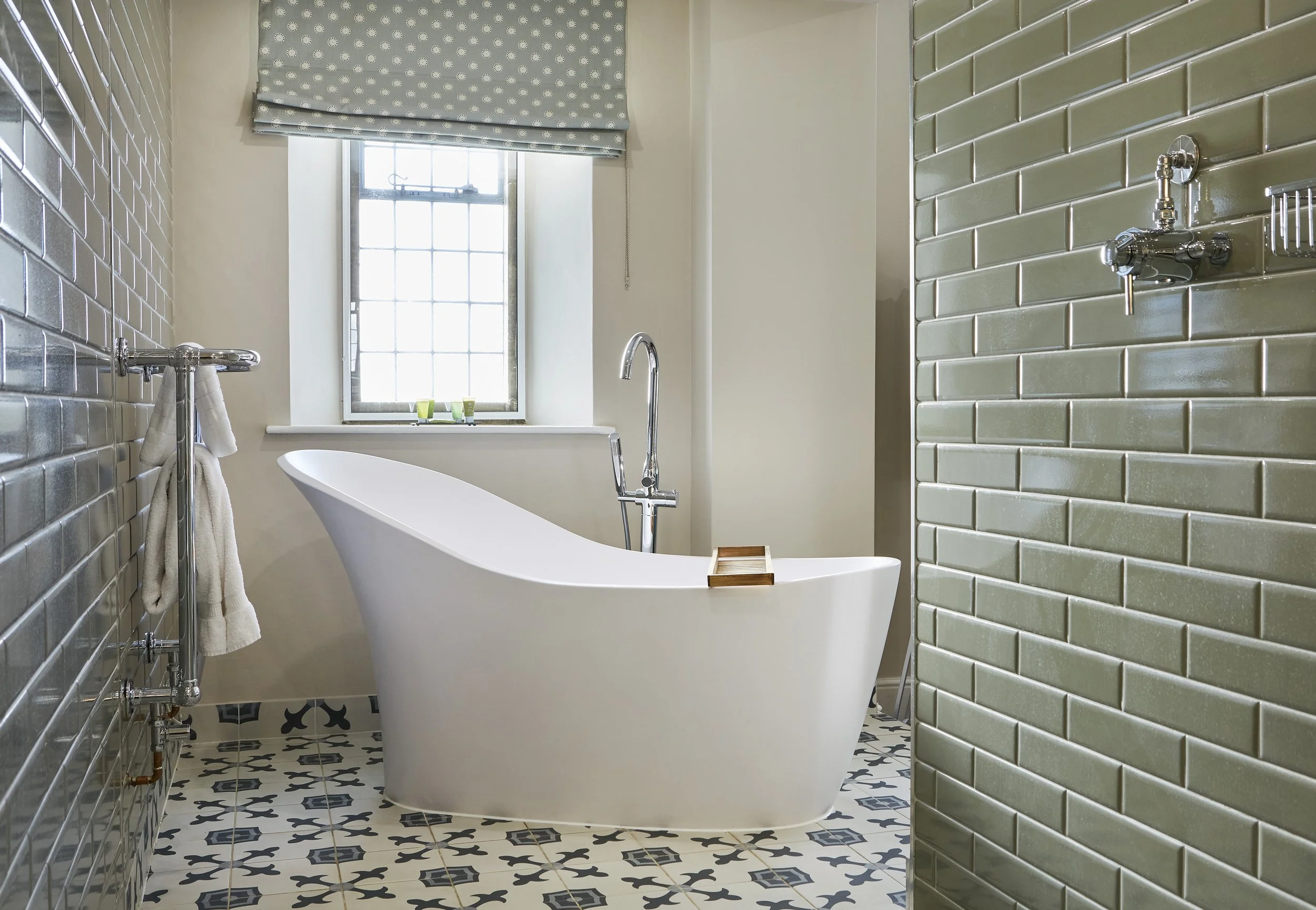Modern bathroom with freestanding white bathtub, patterned floor tiles, green subway tiled walls, and a window with a polka dot Roman shade.