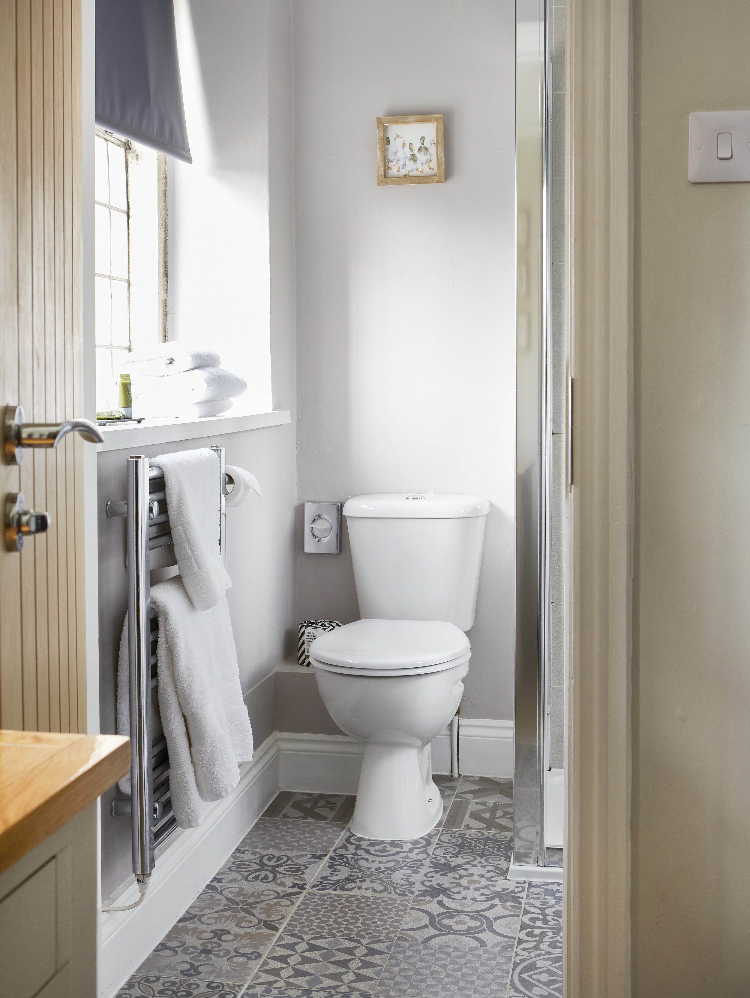 Compact bathroom with toilet, heated towel rack, folded towels, patterned floor tiles, and a wall picture.