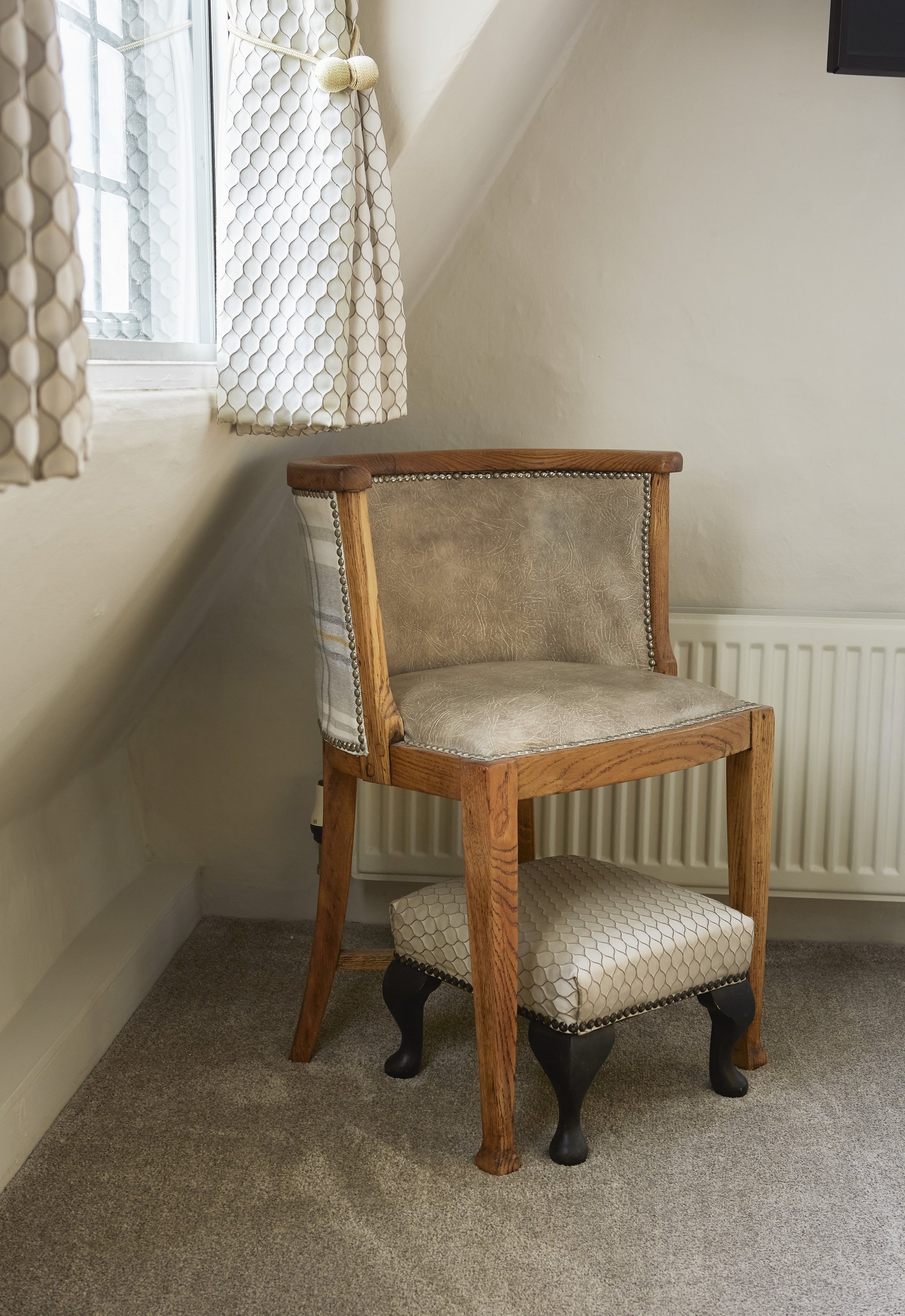 Room corner with a wooden chair and footstool, near a window with patterned curtains.