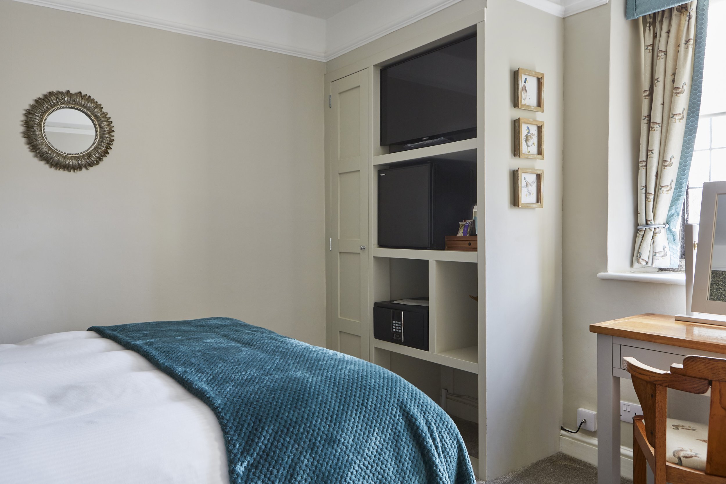 Hotel room with a bed, blue blanket, wall-mounted TV, mini fridge, small safe, mirror, and a window with patterned curtains.