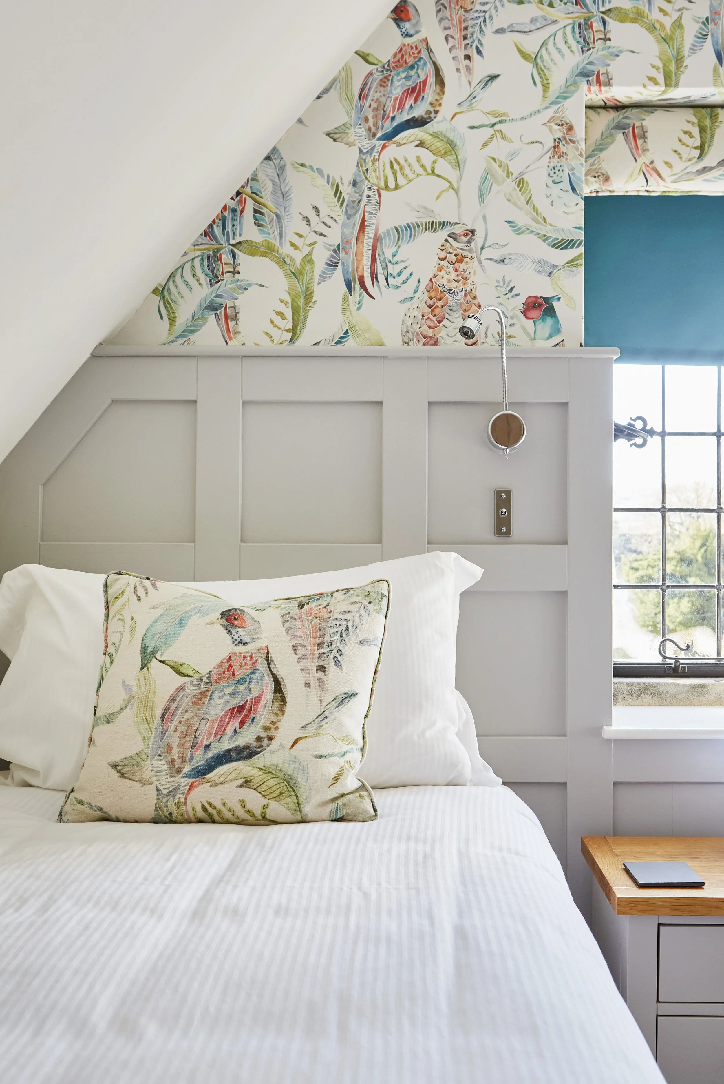 Cozy bedroom with bird-themed decor, featuring a white bed, bird-print pillow, and wallpaper, next to a window.