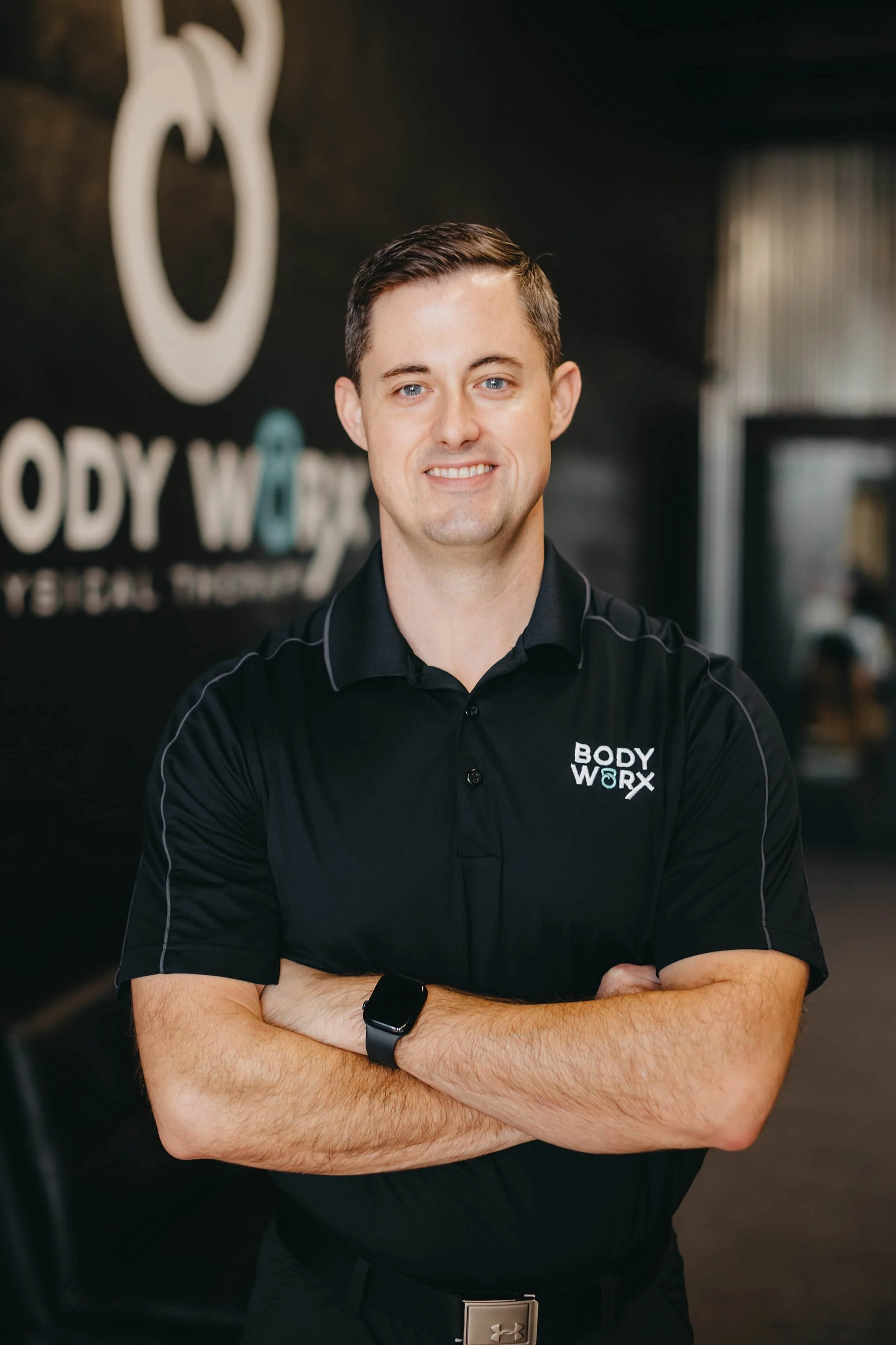 A man in a black BODY WORX shirt stands with arms crossed, smiling in a fitness center with a black background.
