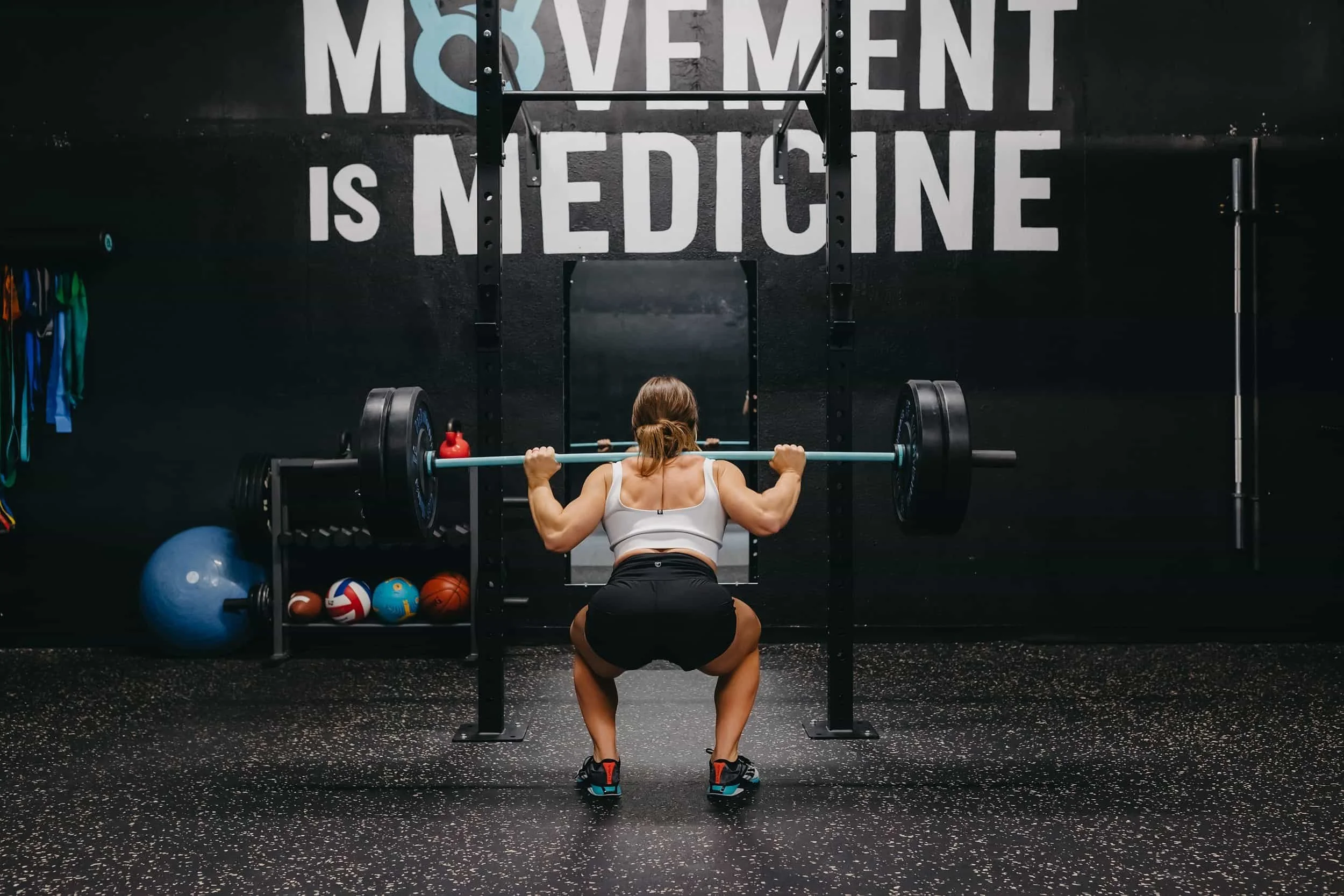A woman performs a barbell squat in a gym with the words Movement is Medicine on the wall behind her.