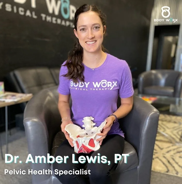 Woman in a purple Body Worx shirt sits in a chair, smiling and holding a pelvic bone model in a physical therapy office.