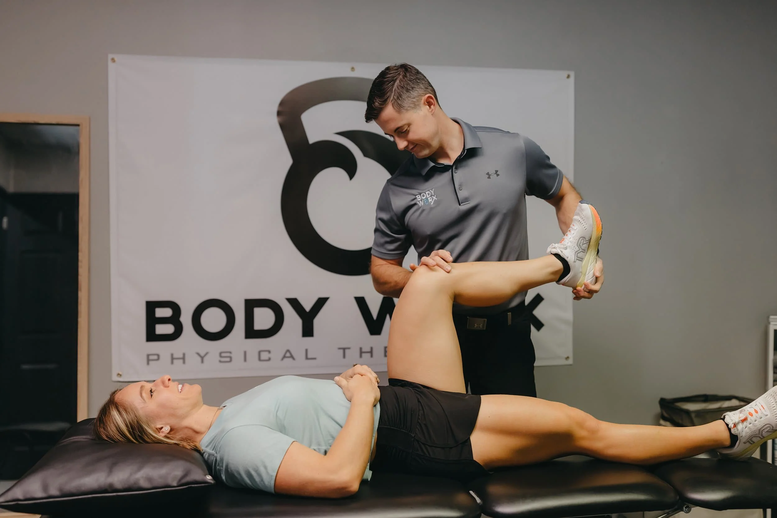 A physical therapist stretches a womans leg on a treatment table at a clinic with a Body Work Physical Therapy sign in the background.