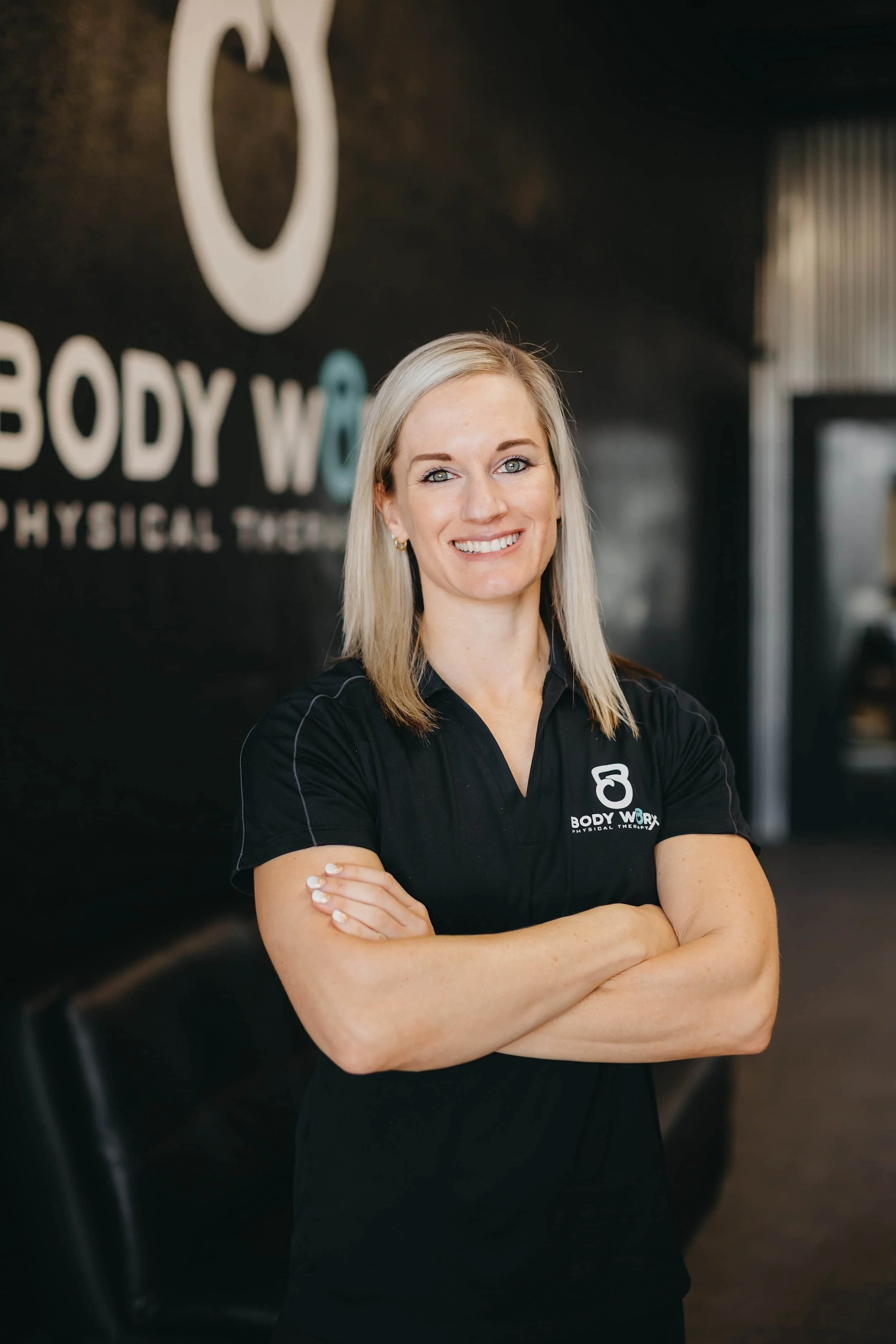 Woman with blonde hair in a black polo shirt stands smiling with arms crossed in a physical therapy clinic.