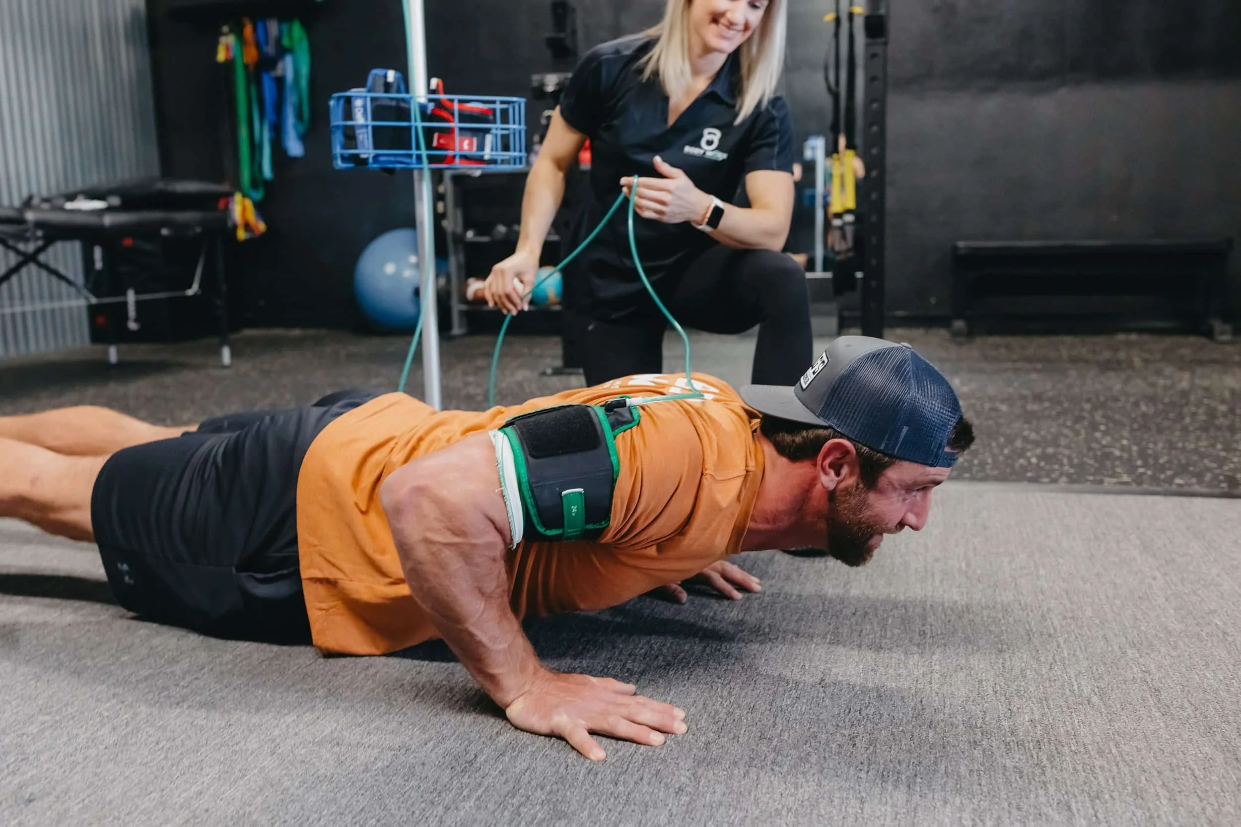 Man doing push-ups with resistance bands while a woman assists in a gym setting.
