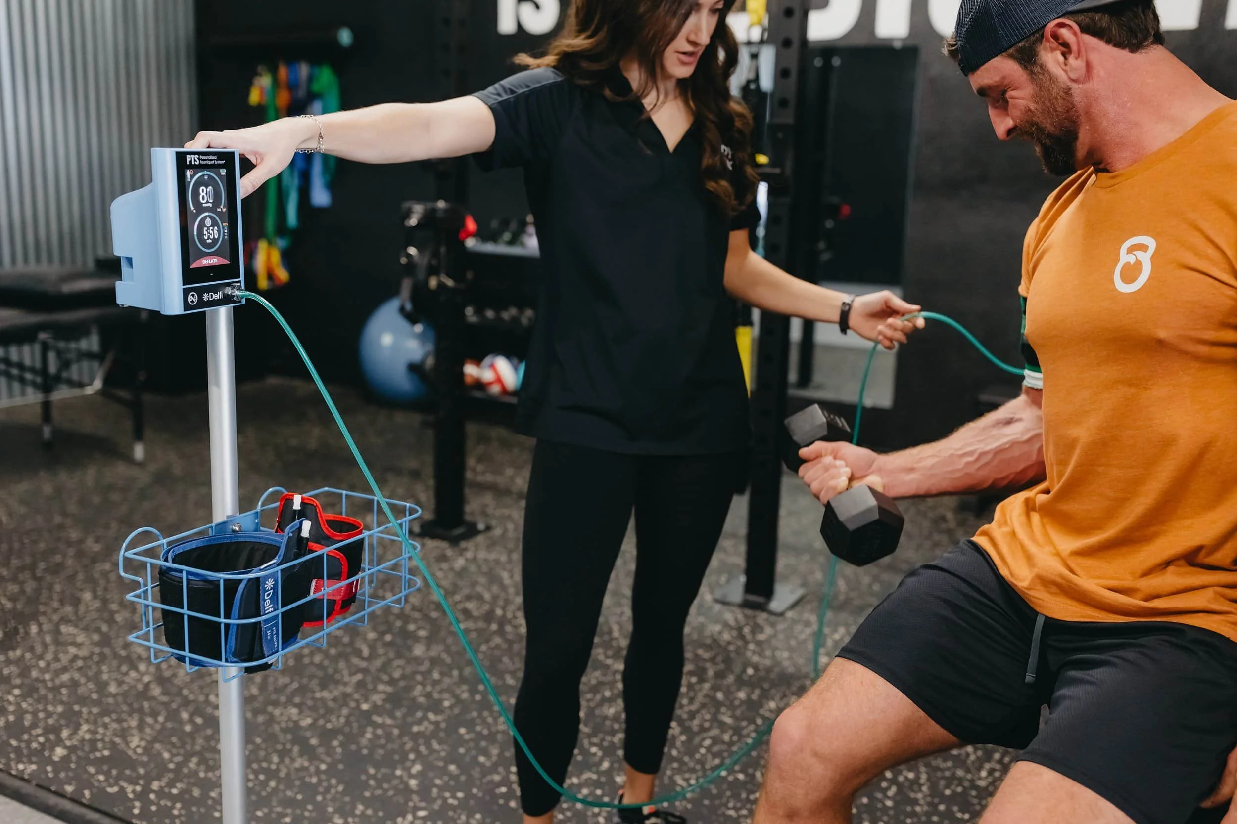A man lifts a dumbbell while a woman in black scrubs checks equipment attached to his arm in a gym setting.