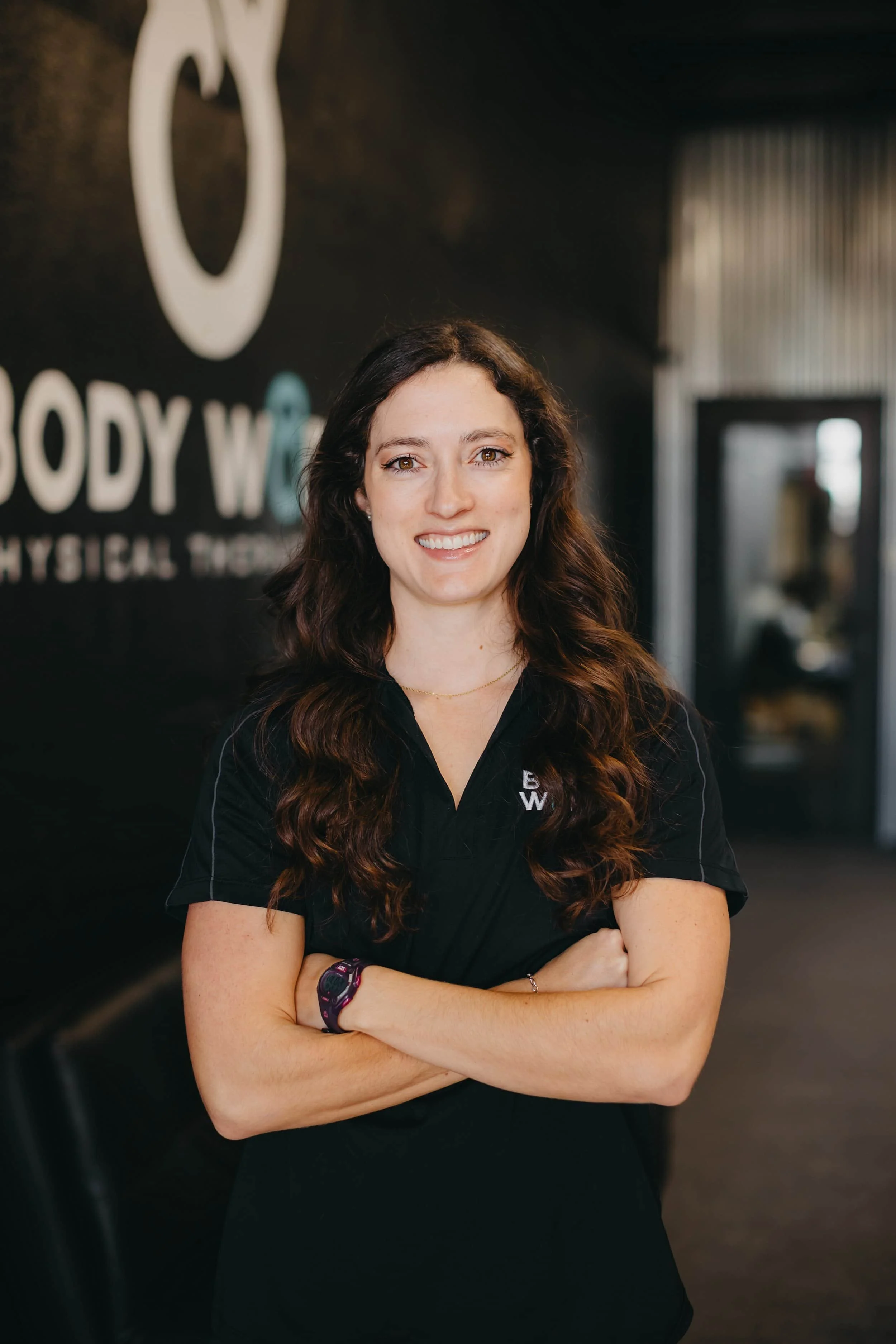 Woman with long wavy hair in a black shirt stands smiling with arms crossed in a physical therapy clinic.
