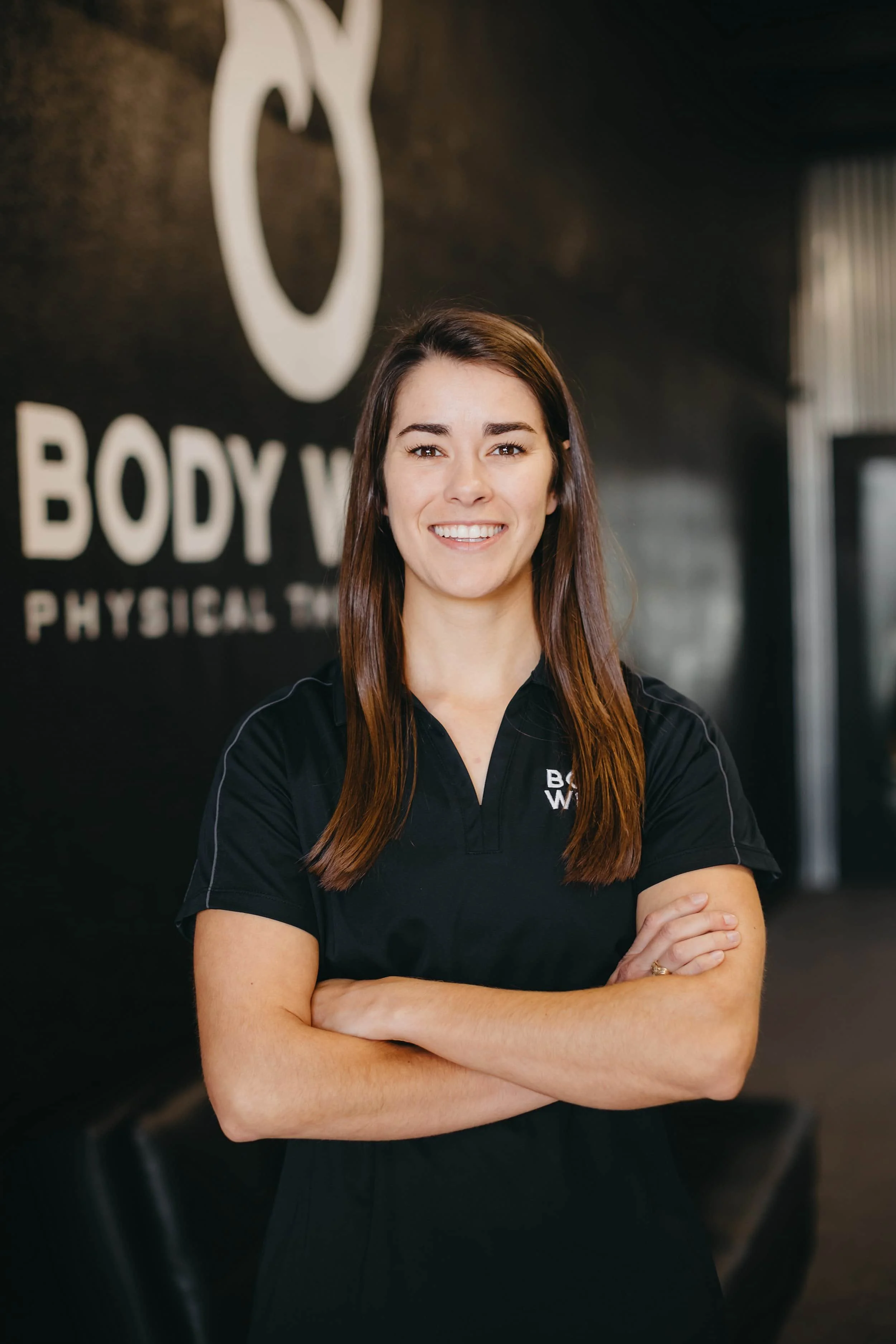 A woman in a black polo shirt stands with arms crossed, smiling in front of a Body Works Physical Therapy sign.