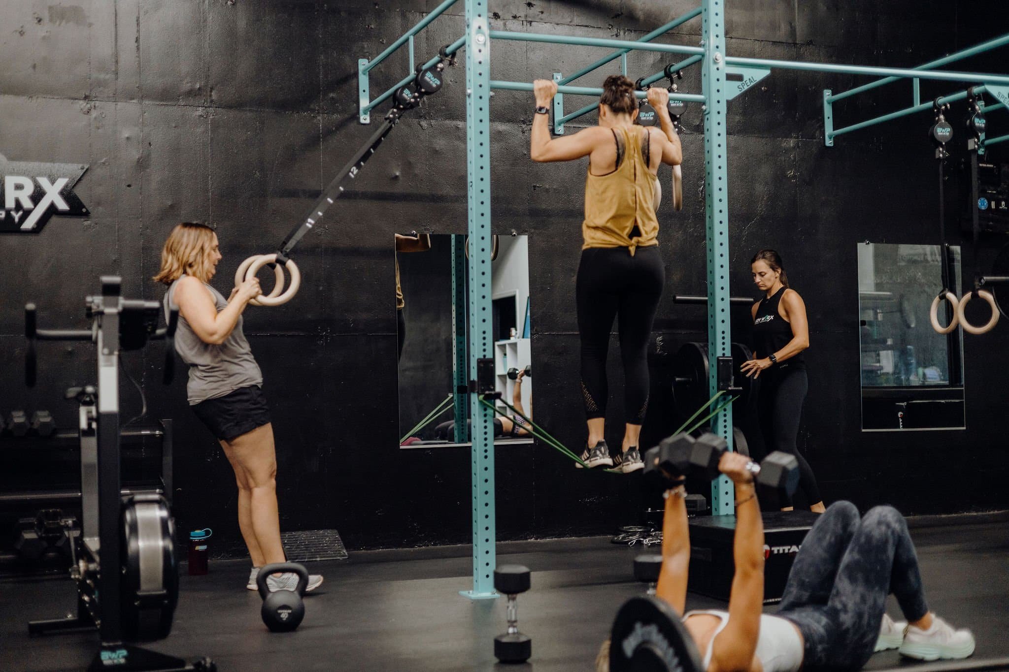 Four women work out in a gym, using pull-up bars, gymnastic rings, dumbbells, and a rowing machine.