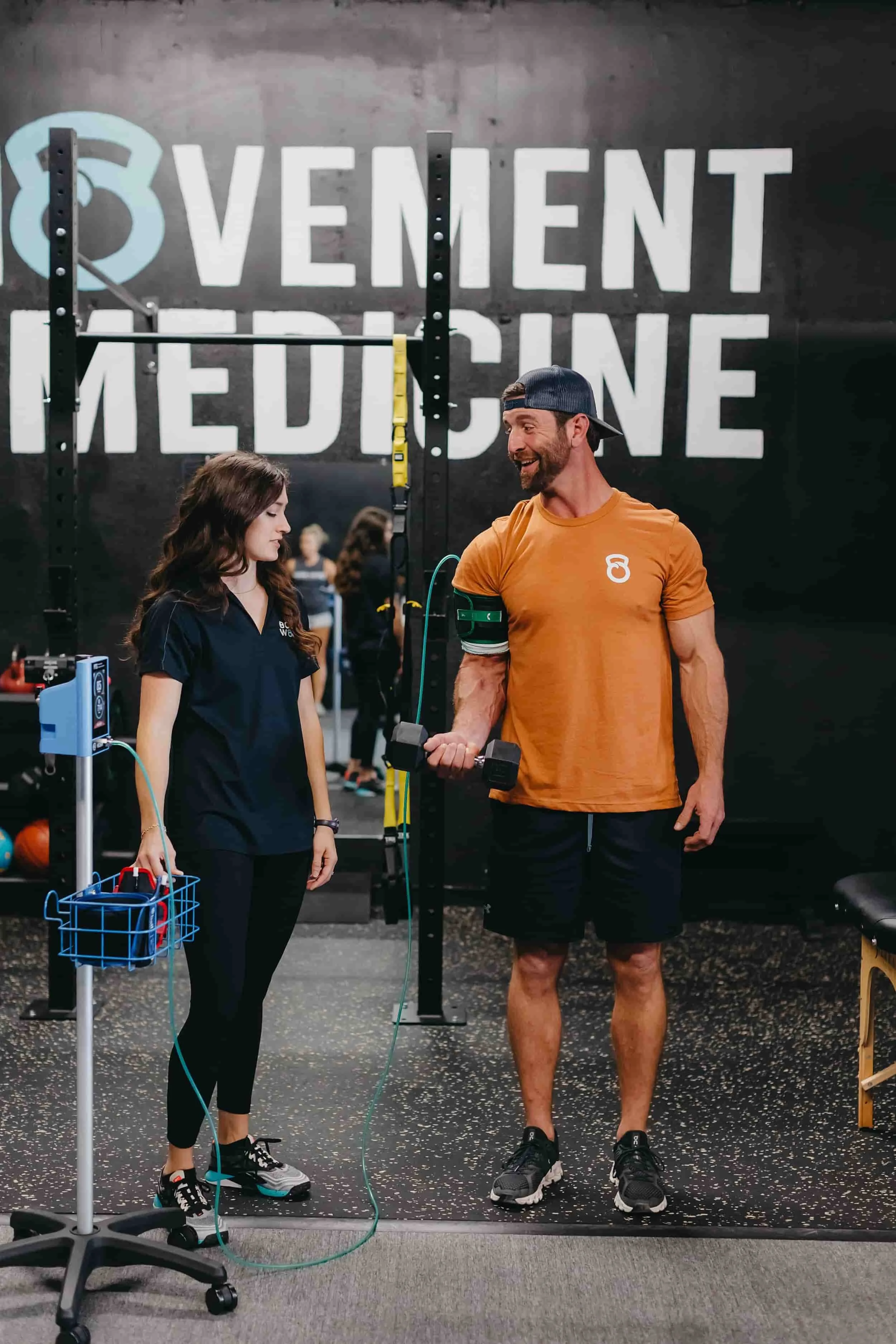 A man lifts a dumbbell with a blood flow restriction band as a woman in scrubs observes in a gym labeled Movement Medicine.