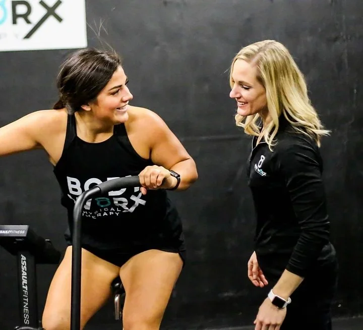 Two women at a gym, one exercising on a stationary bike while the other stands beside her, both smiling.
