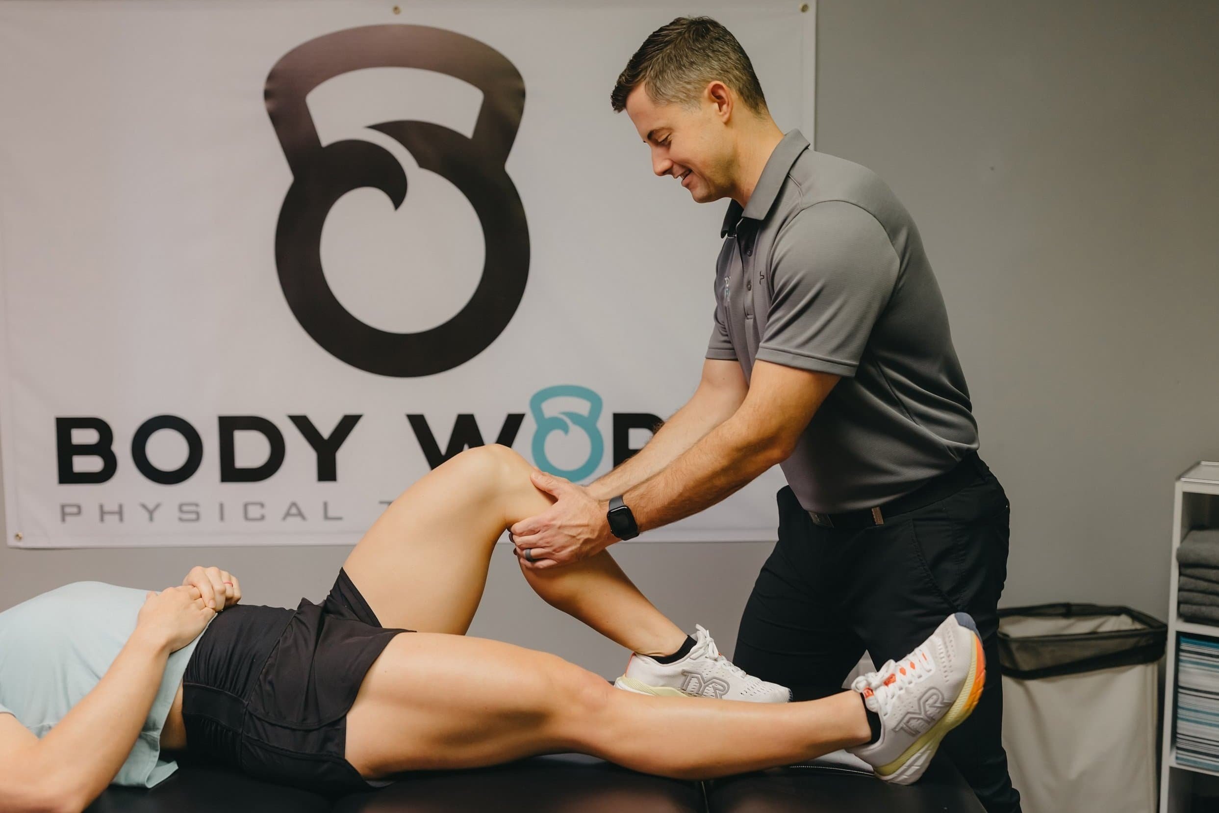 A physical therapist assists a patient with a leg exercise in a clinic, beside a Body Work Physical banner.