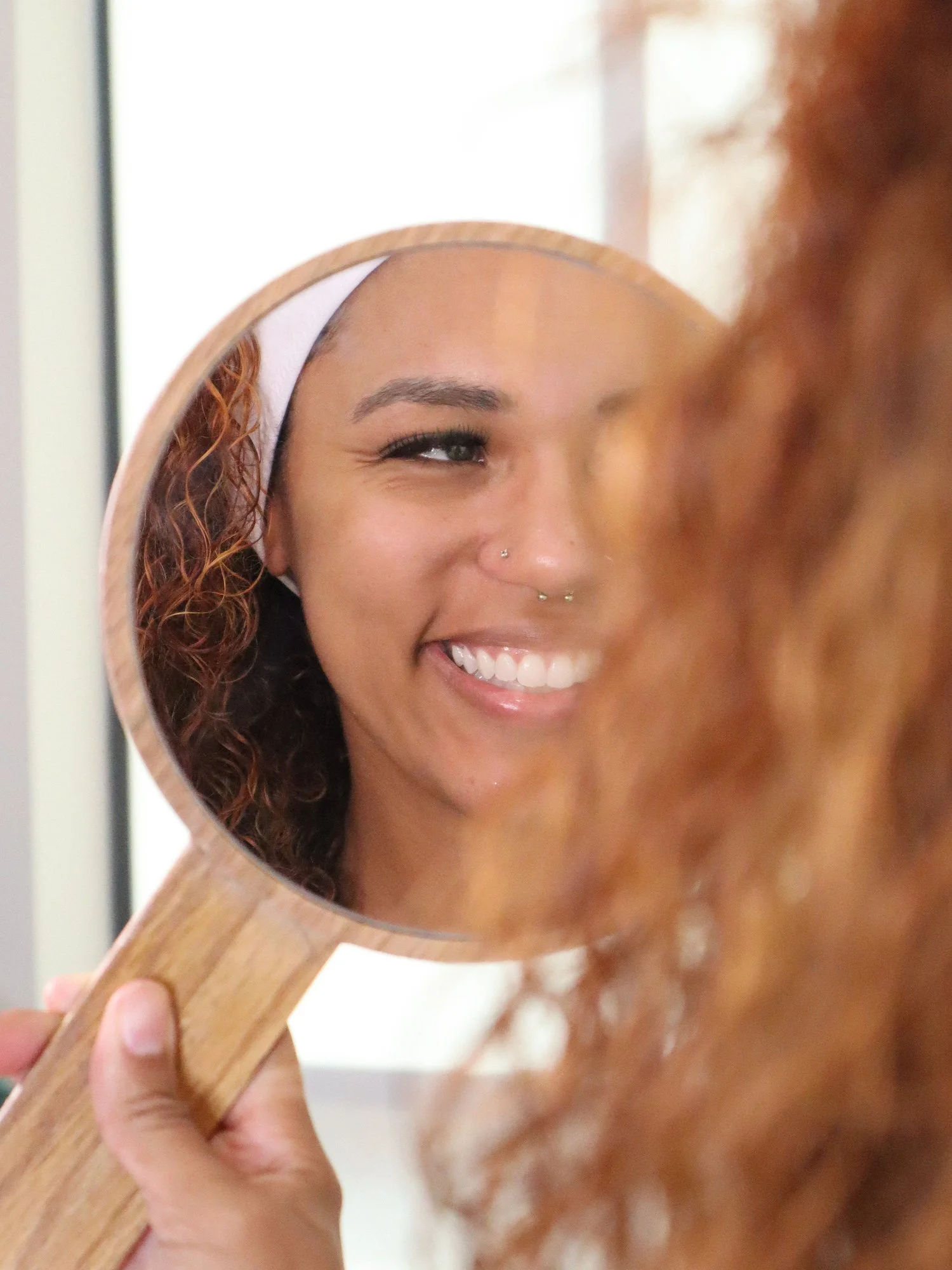 A woman smiling and looking at her reflection in a handheld mirror, which shows her face with a happy expression, curly hair, and a nose piercing.