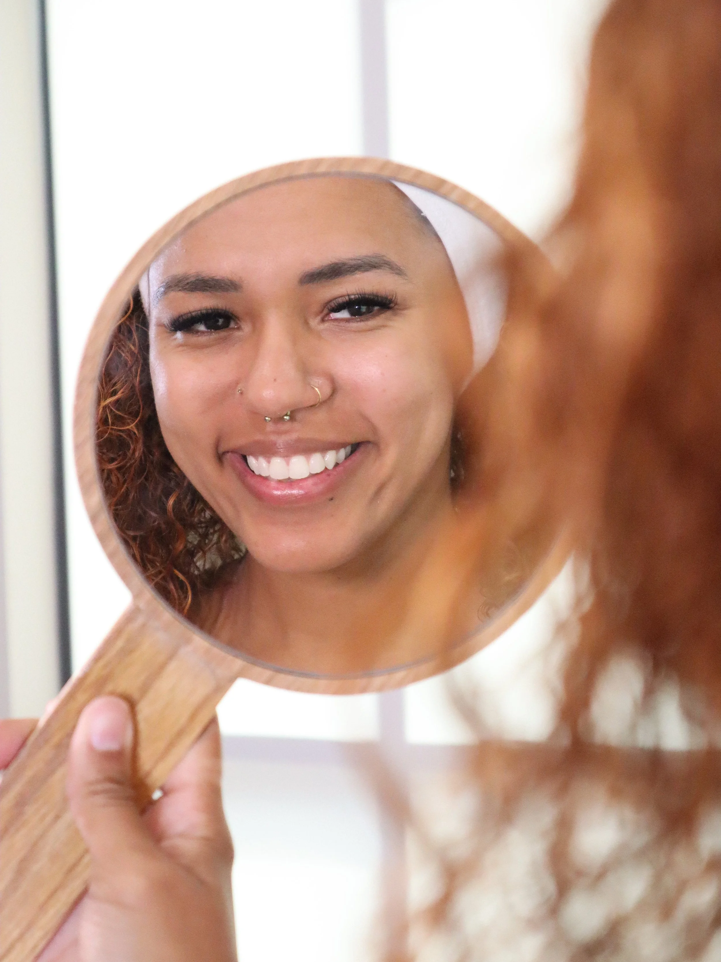 Person holding a mirror reflecting their smiling face with curly hair and multiple facial piercings.
