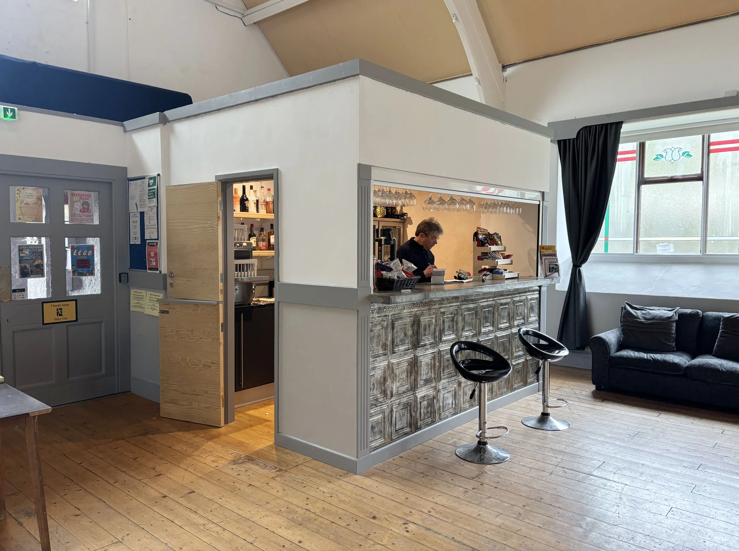 A bar decorated with colorful bunting and balloons, with three people standing at the bar counter engaged in conversation.
