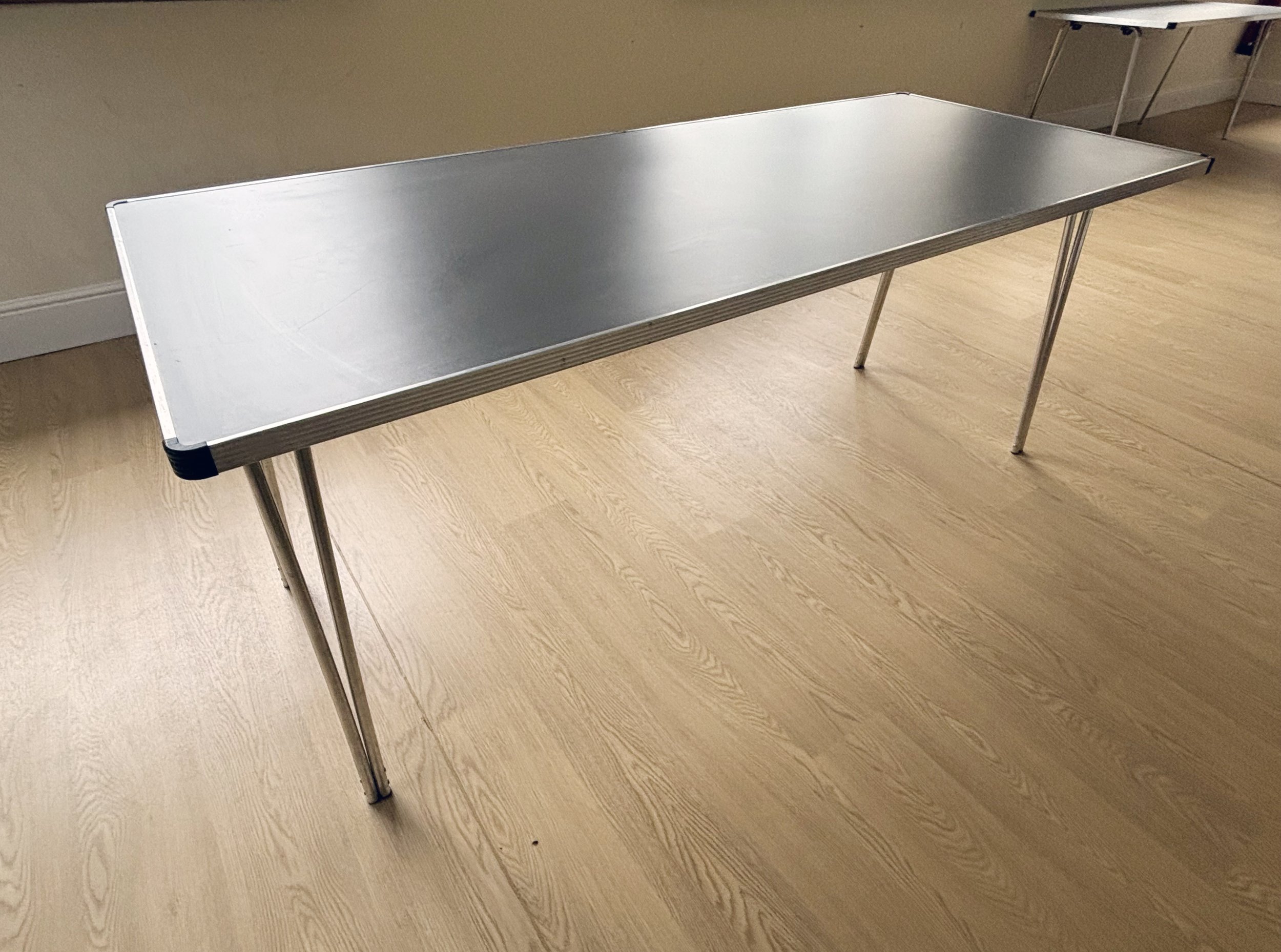 A beige folding table with black metal legs, set against a white wall with a gray trim on a wooden floor.