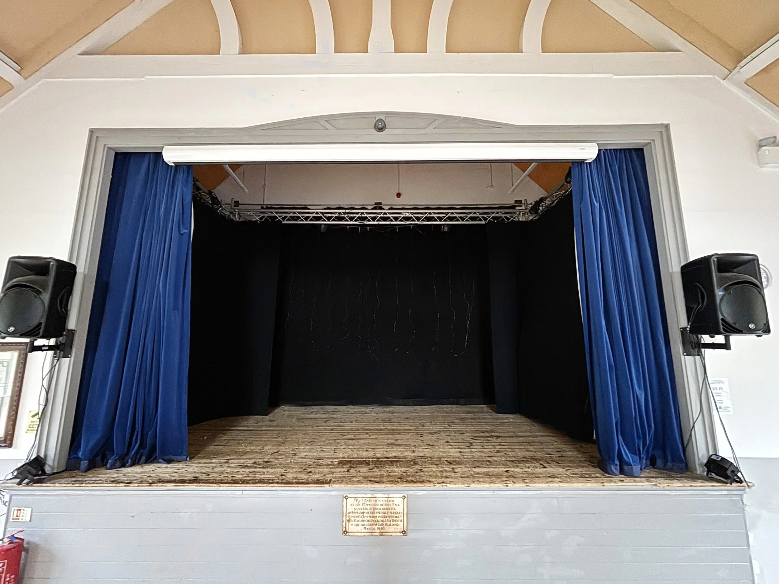 An empty multi-purpose hall with a wooden floor, stage area with stacked chairs, and walls with small windows and framed pictures or decorations.