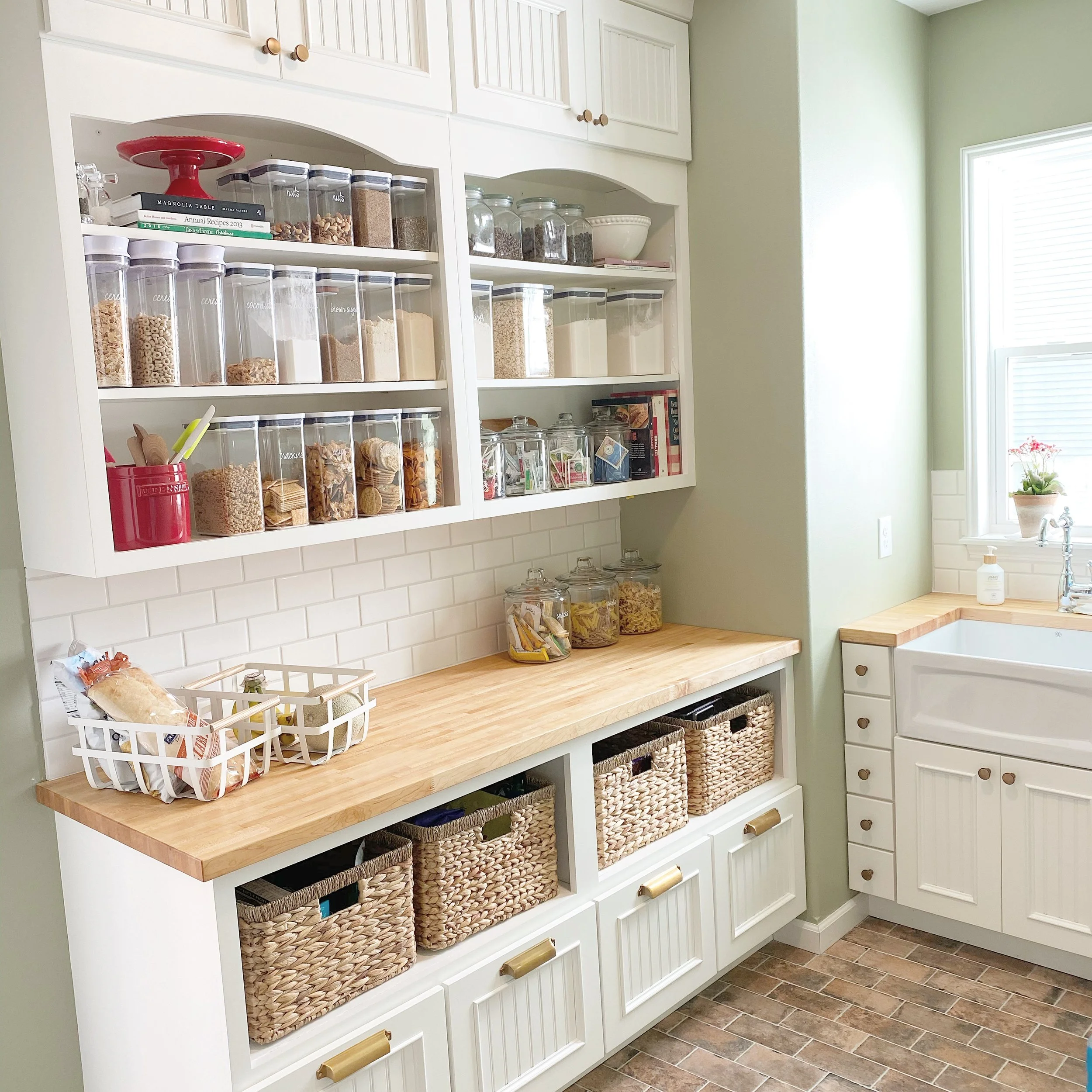 A well-organized kitchen pantry area with open white cabinets filled with labeled containers of dry goods, a wooden countertop with jars of snacks, and woven baskets for storage, adjacent to a white sink under a window with a potted plant.