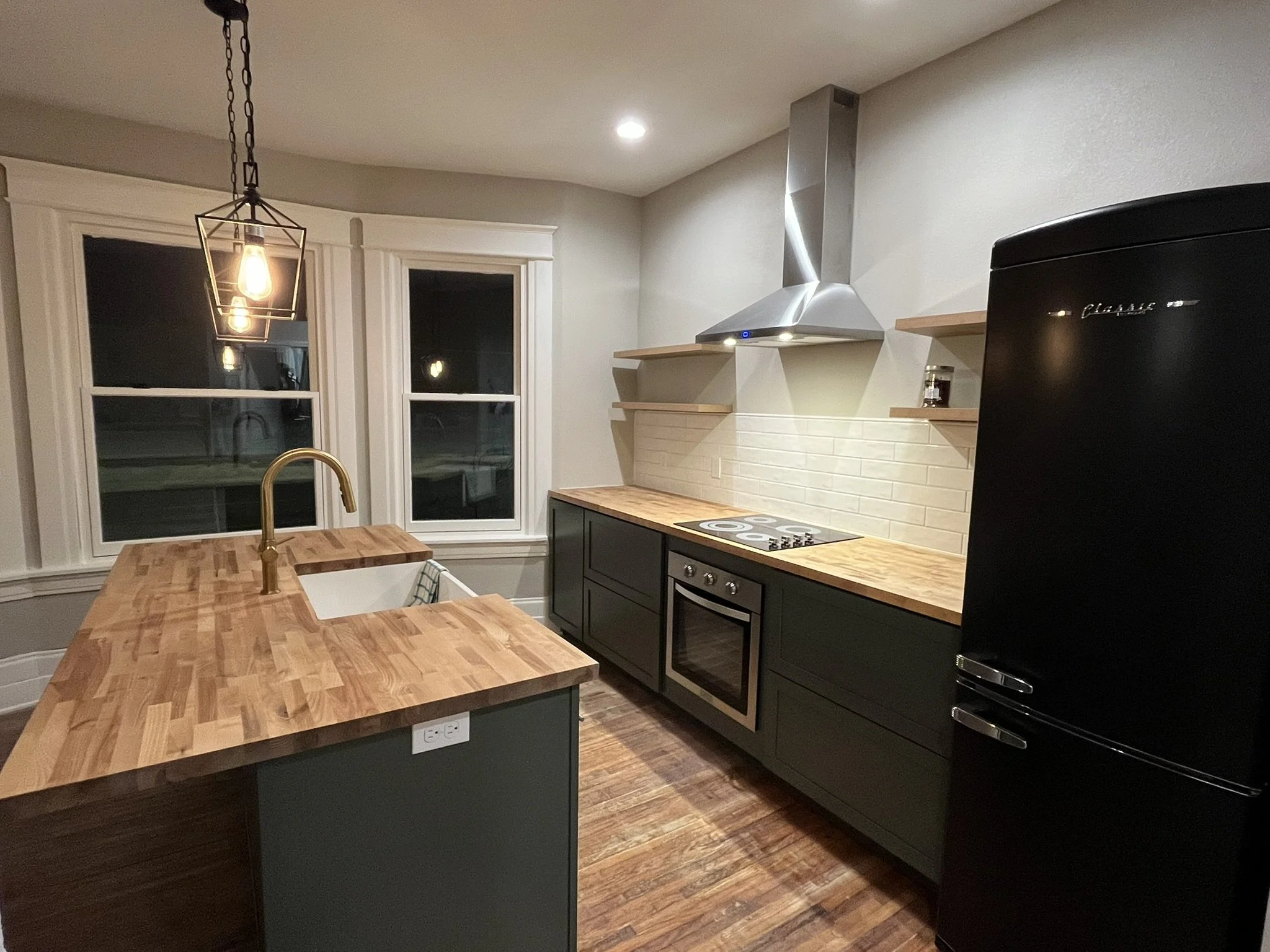 Modern kitchen with dark green cabinets, wooden countertops, two large windows, a black refrigerator, a stainless steel range hood, and an hanging pendant light over a kitchen island.
