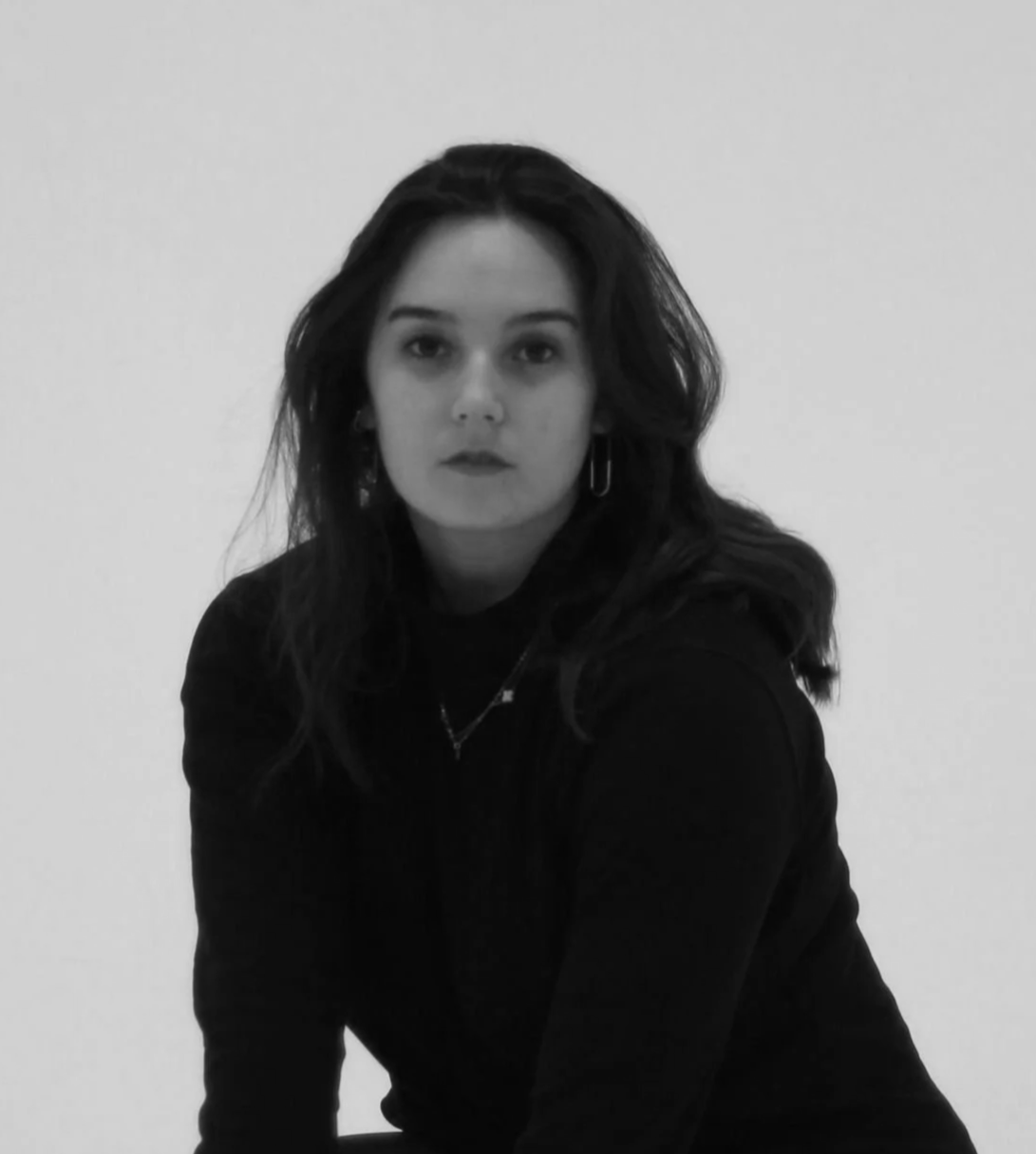 Black and white photo of a young woman with dark hair, wearing a dark top and earrings, looking directly at the camera against a plain light background.