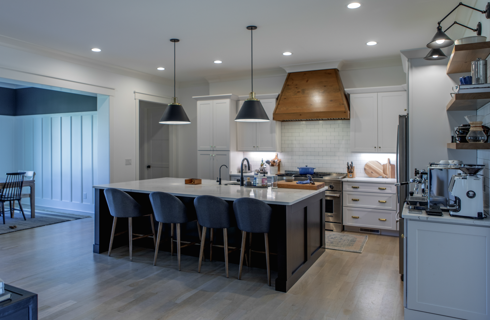 Modern kitchen with white cabinets, a large island with black base and four blue upholstered bar stools, pendant lights, and stainless steel appliances. Open shelving and a wooden range hood over a stove.
