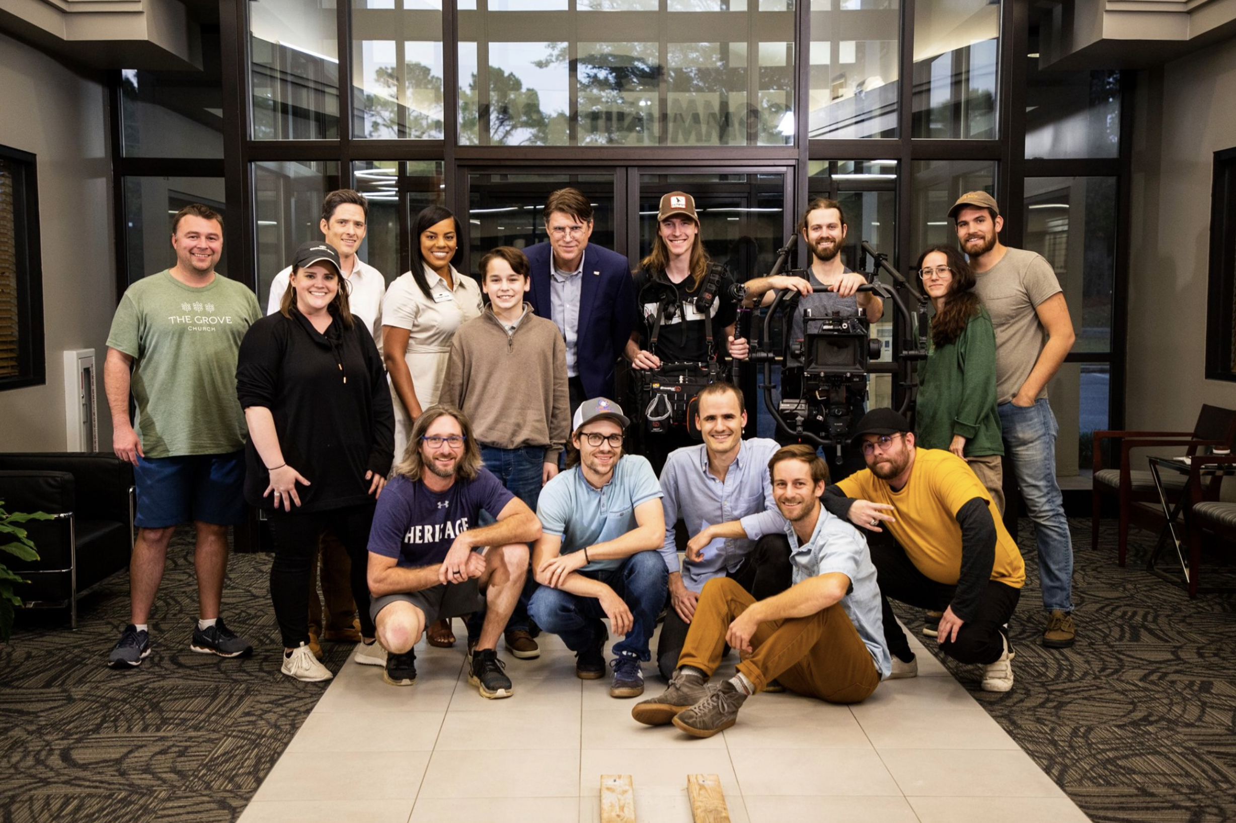 A group of 15 people, including men and women of various ages and ethnicities, pose for a photo indoors near a large window with a camera crew and equipment. Some are standing, some are kneeling, and they all seem happy.