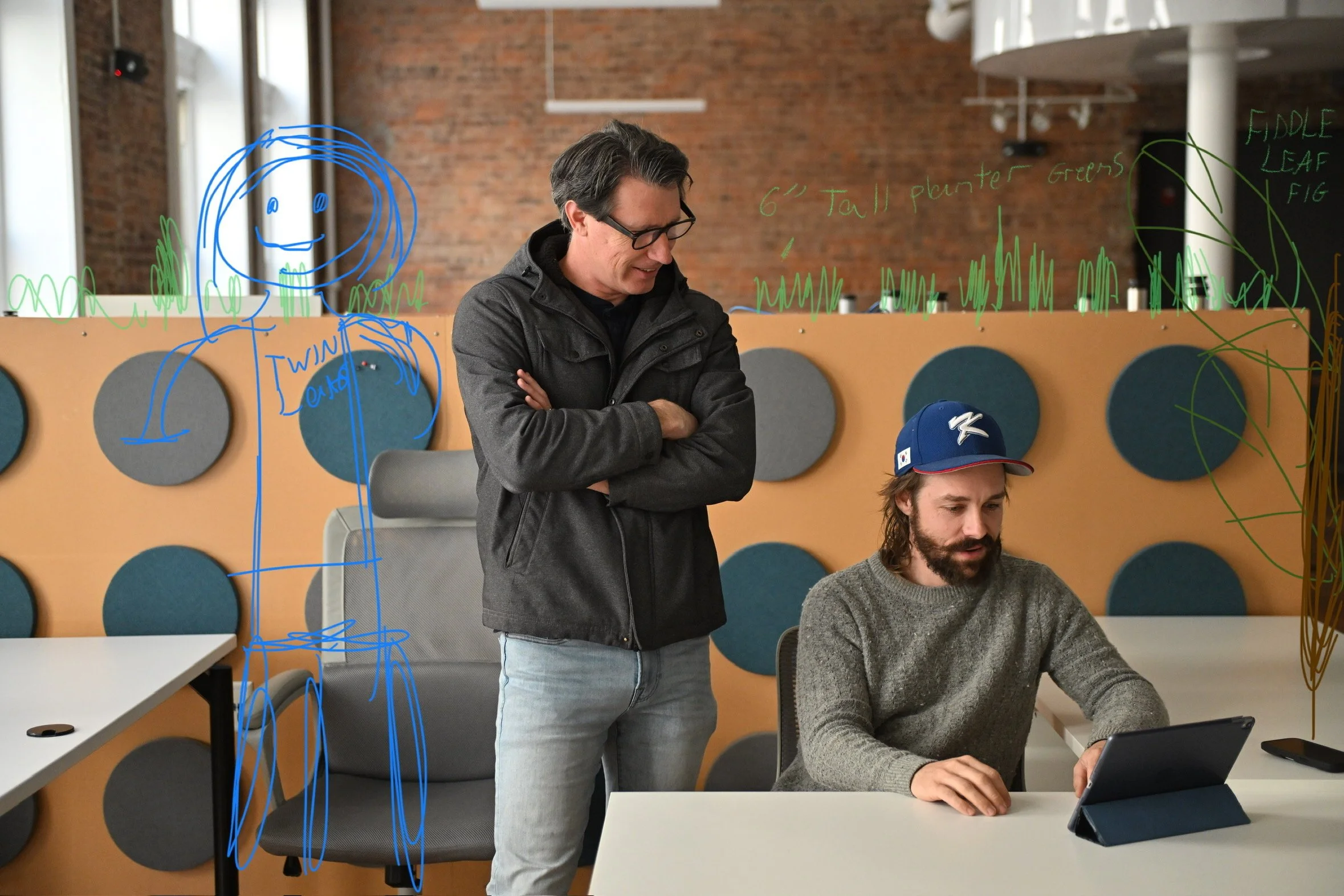 Two men in an office, one standing and the other sitting, with colorful drawings and notes on a glass wall behind them. The sitting man is using a tablet, and the standing man is observing.