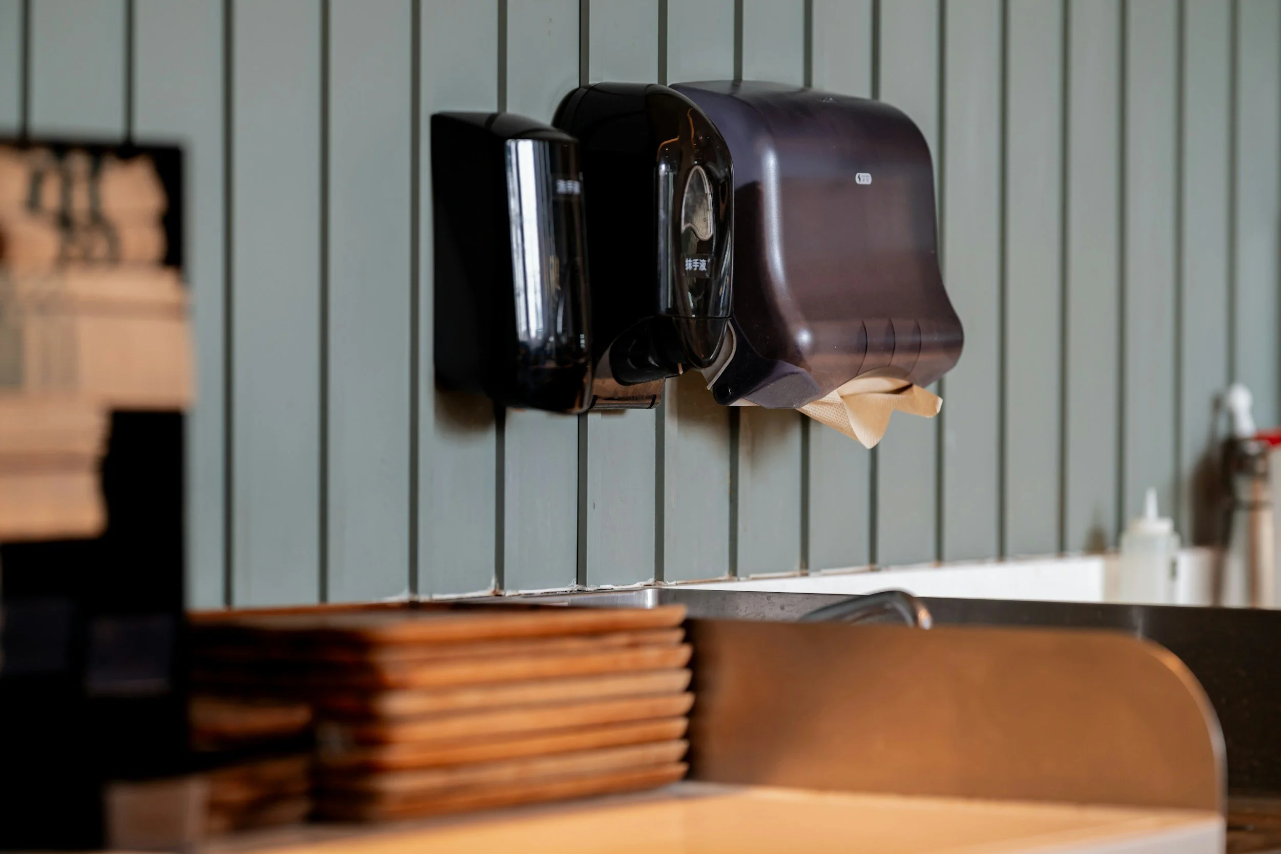 Wall-mounted black paper towel dispenser and napkin holder in a restaurant or cafe setting with stacked wooden plates below.