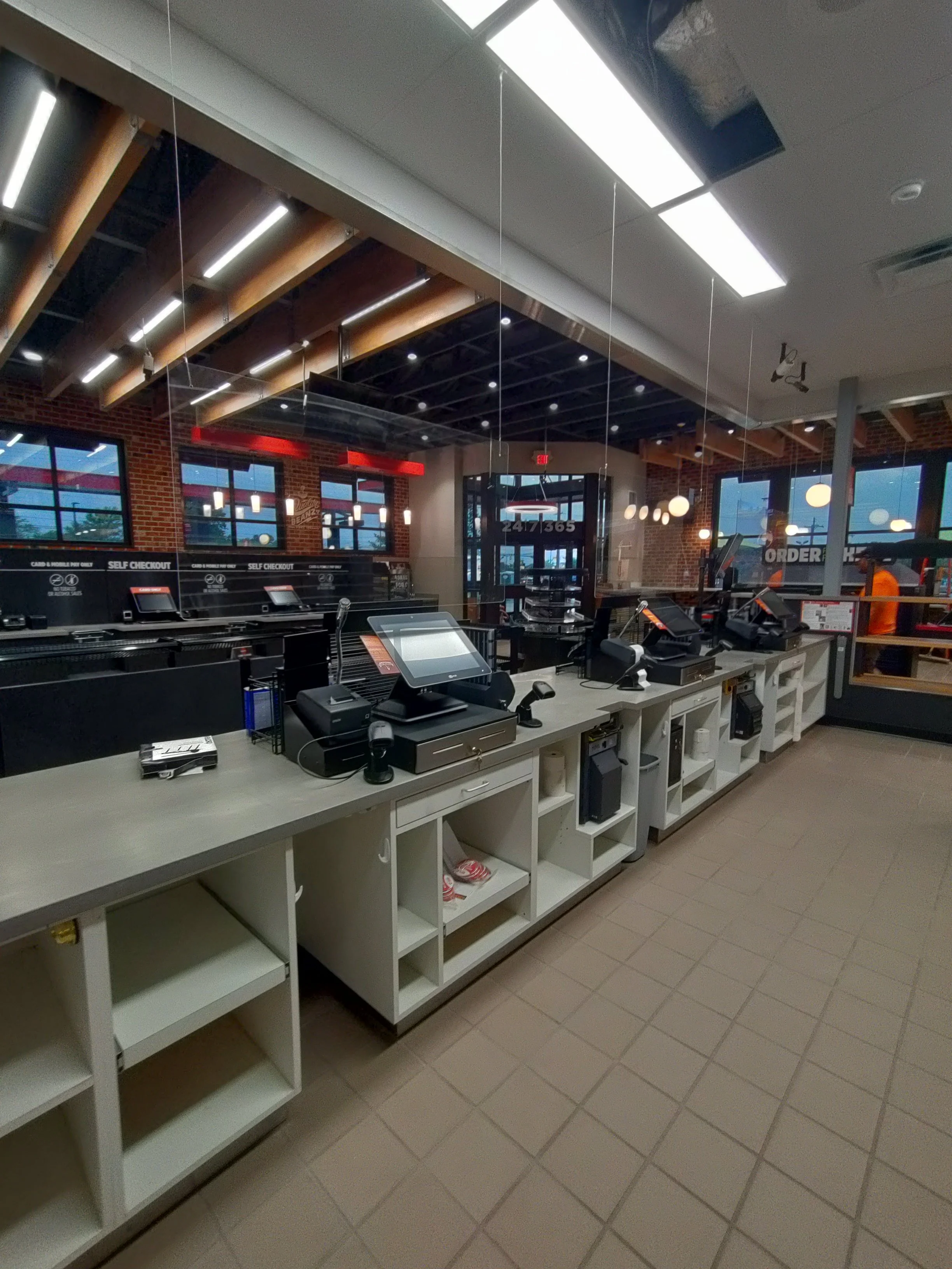 Inside a modern fast-food restaurant with checkout counters, self-service kiosks, and a visible order pickup area. The background features large windows, brick walls, and ceiling lights.