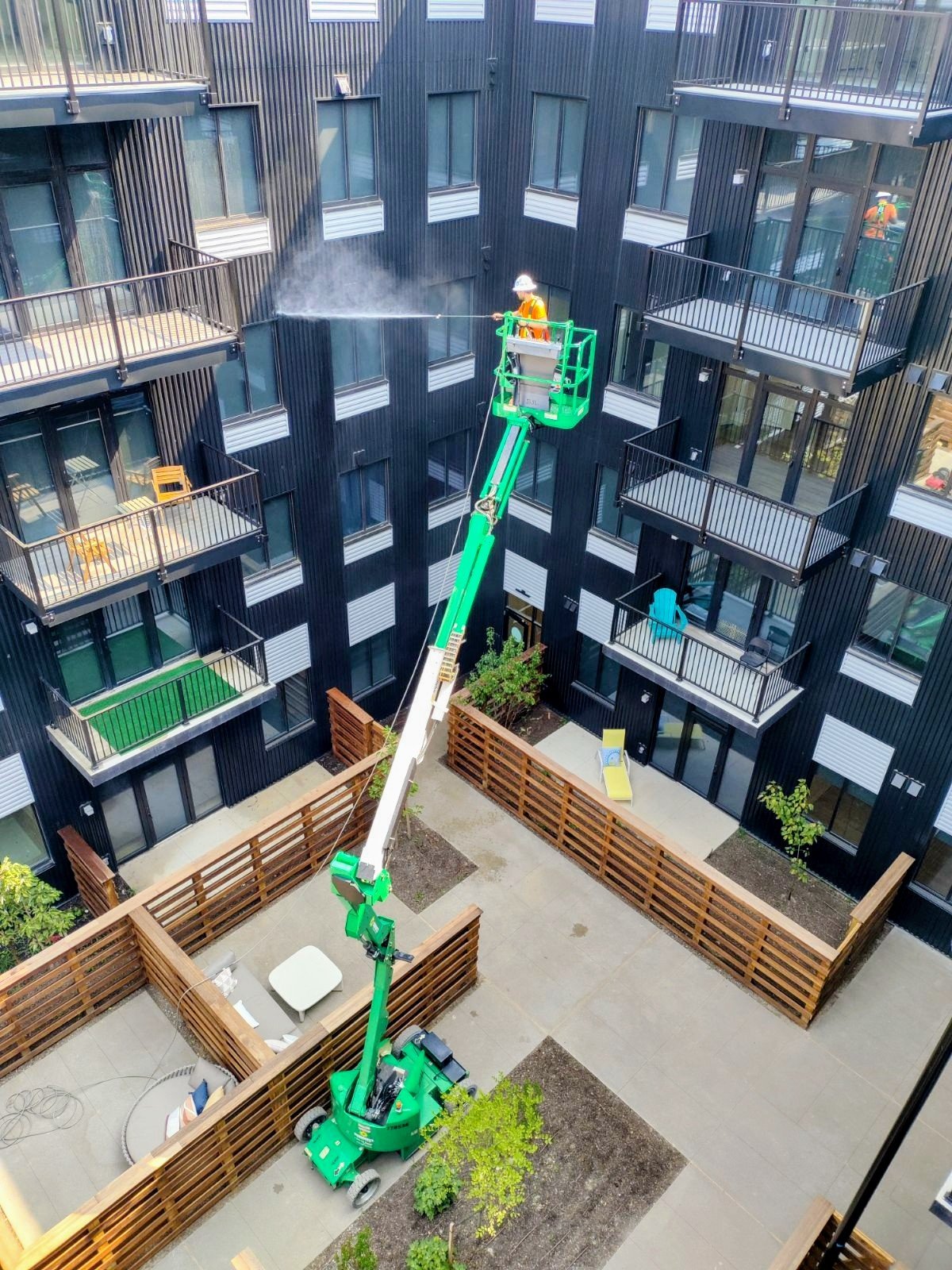 A man wearing a blue shirt, gray overalls, a dark green cap, and glasses is power washing a brick wall on a sunny day with a blue sky. He is standing outdoors, holding a pressure washer spray wand, with a small gray utility cart nearby.