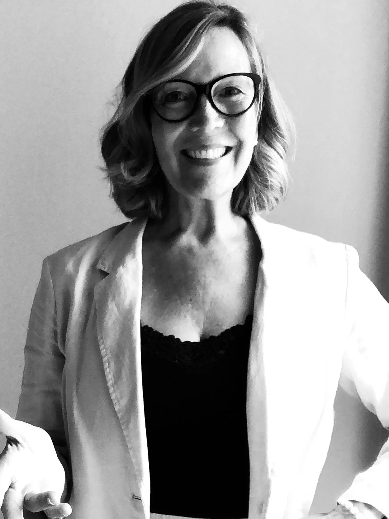 Black-and-white photo of a smiling woman with wavy hair, glasses, wearing a blazer and a lace-trimmed top.