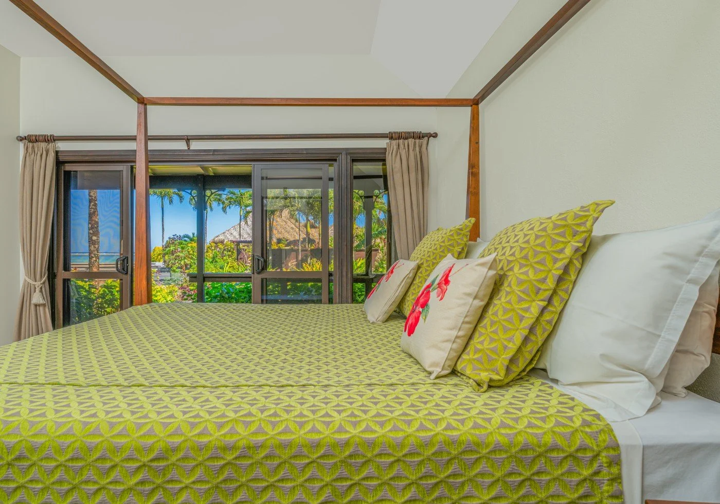 Hotel room with a bed, yellow patterned bedding, decorative pillows with pink floral designs, large window with view of tropical trees, mountains, and ocean in the background.