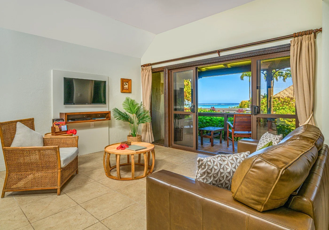 Living room in a tropical resort with sliding glass doors leading to a balcony with lagoon view, surrounded by lush green foliage and palm trees. The room has wicker and leather furniture, a flat-screen TV, and a small round coffee table.