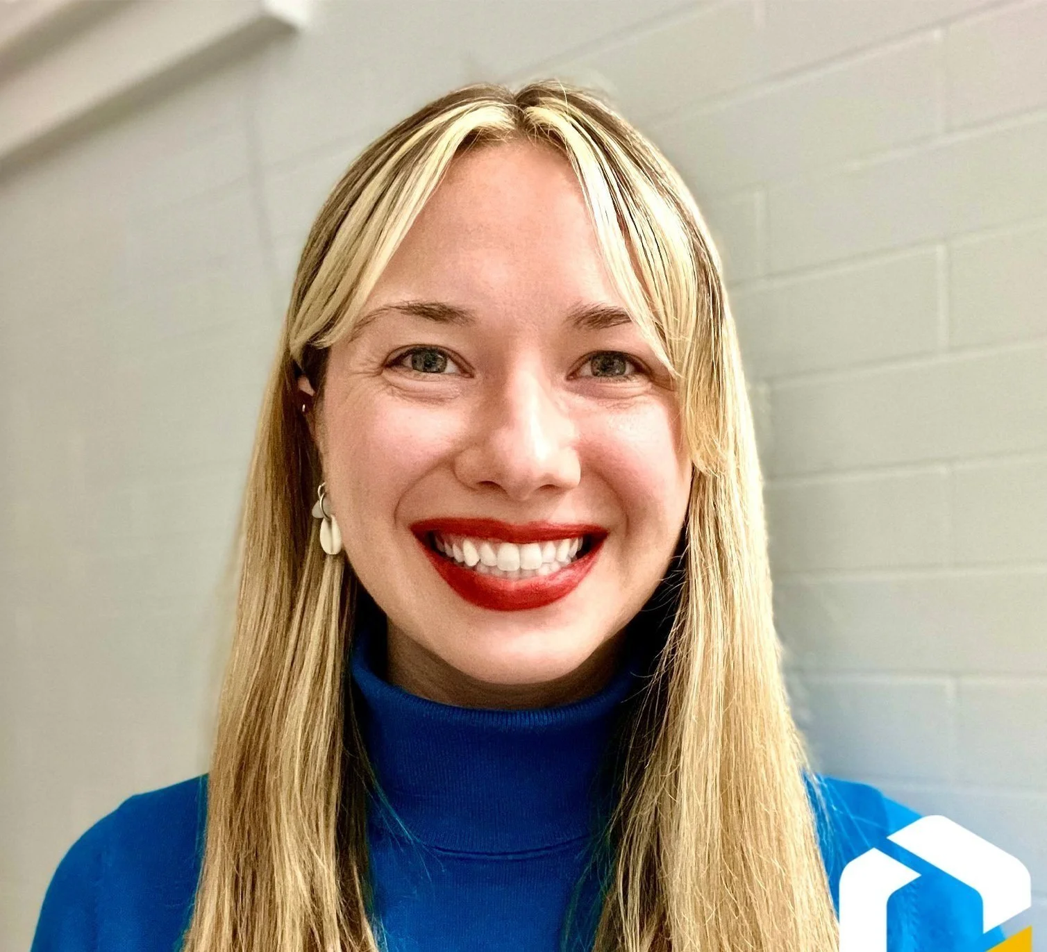 Close-up of a smiling woman with blonde hair, wearing a blue turtleneck, earrings, and red lipstick, standing against a light-colored brick wall.