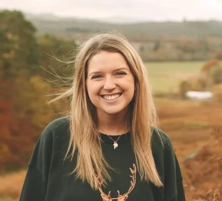 A young woman with long blonde hair smiling outdoors, with a scenic landscape of trees and fields in the background.