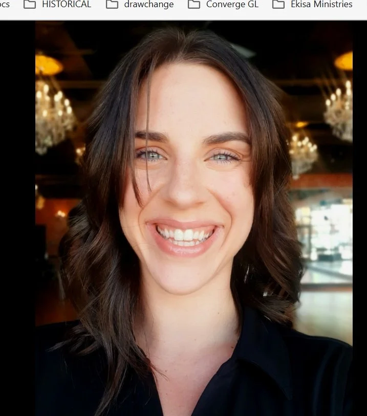 Close-up of a smiling woman with light skin, shoulder-length wavy brown hair, and bright eyes, indoors with chandeliers in the background.