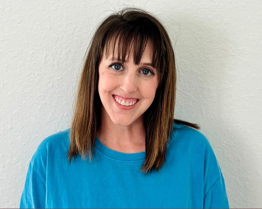 Smiling woman with shoulder-length brown hair wearing a blue shirt standing against a plain white wall.