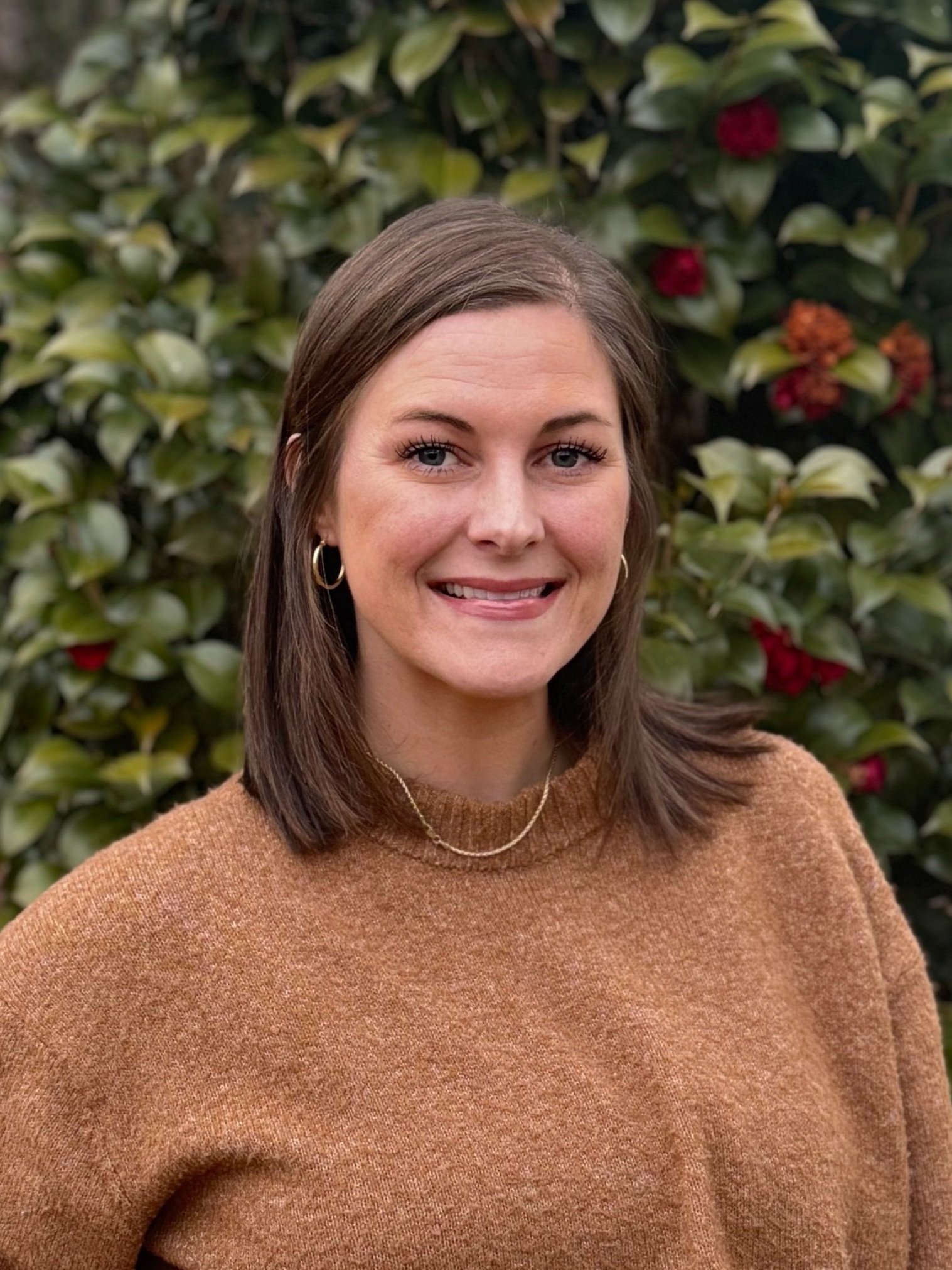 A woman with shoulder-length brown hair, wearing hoops, a gold necklace, and a brown sweater, smiling outdoors with a green leafy background and red flowers.