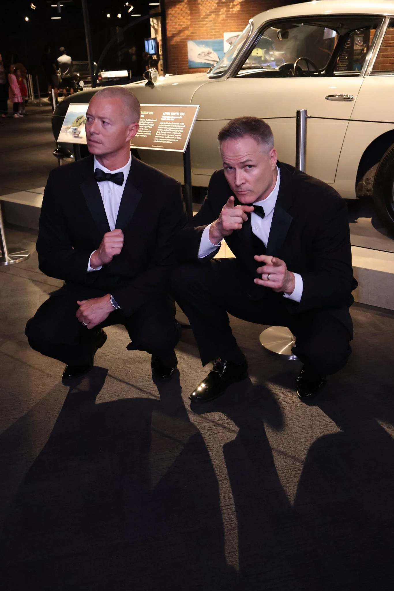 Two men dressed as James Bond in tuxedos squatting in front of a vintage car, with one pointing at the camera, inside a museum exhibit.