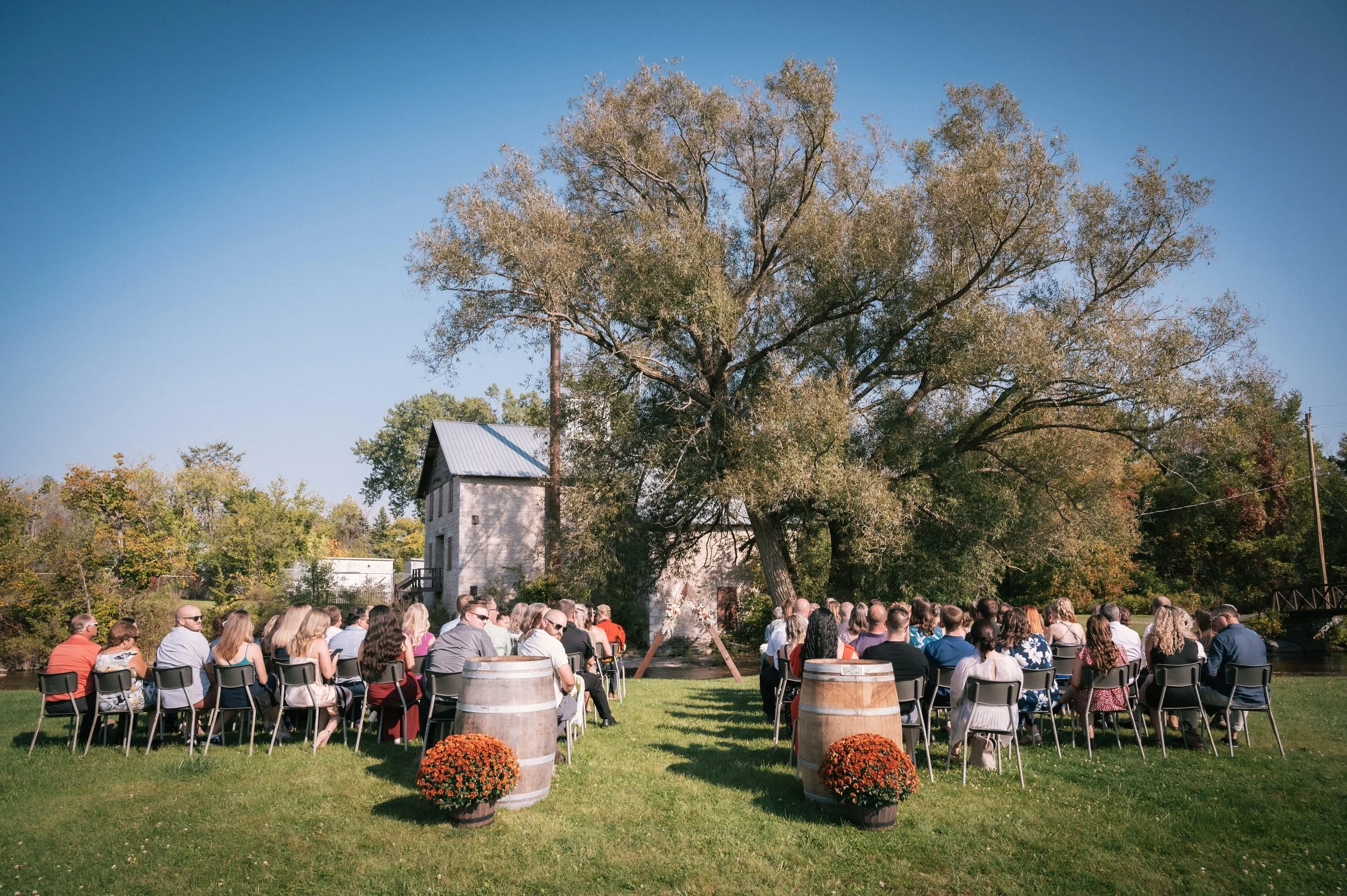 A group of people seated outdoors on chairs under a large tree, likely attending a ceremony or event, with barrels and flowers in the foreground and an old building in the background on a sunny day.