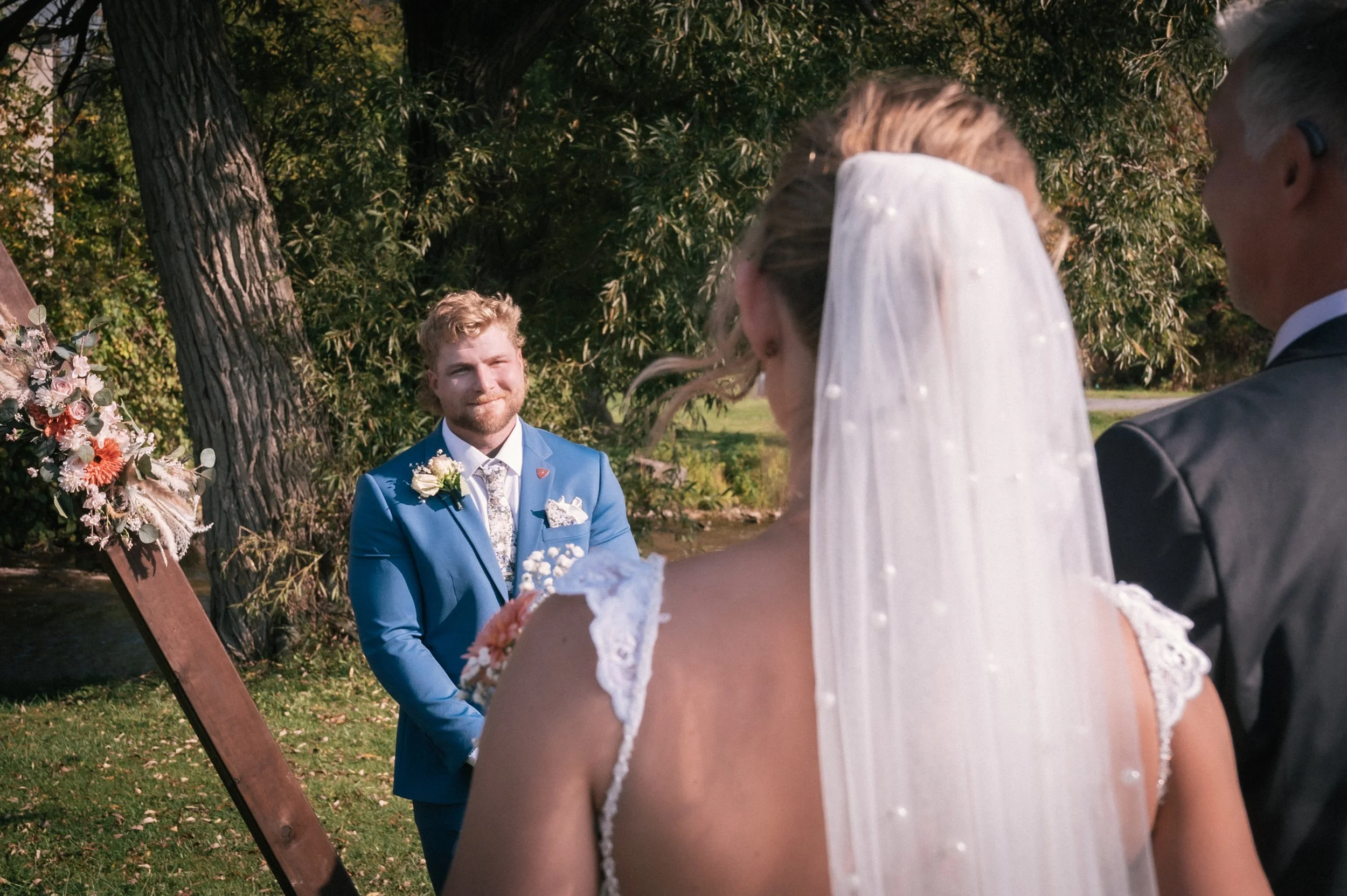 A bride in a white wedding dress and veil, a groom in a gray suit, and a man in a blue suit with a boutonniere, participating in a wedding ceremony outdoors near trees and greenery.