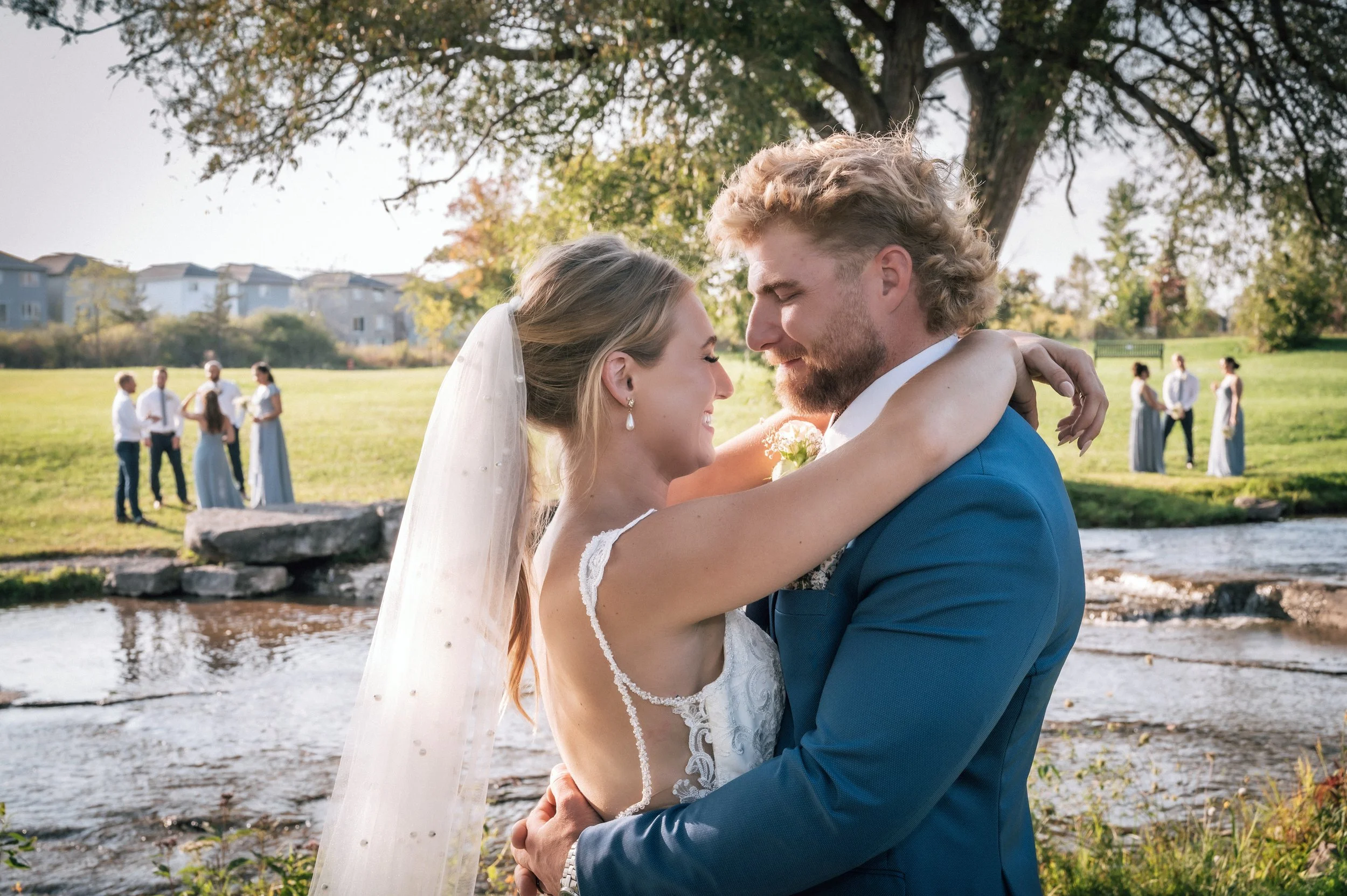 A bride and groom embracing by a river, with a wedding party in the background in a park setting.