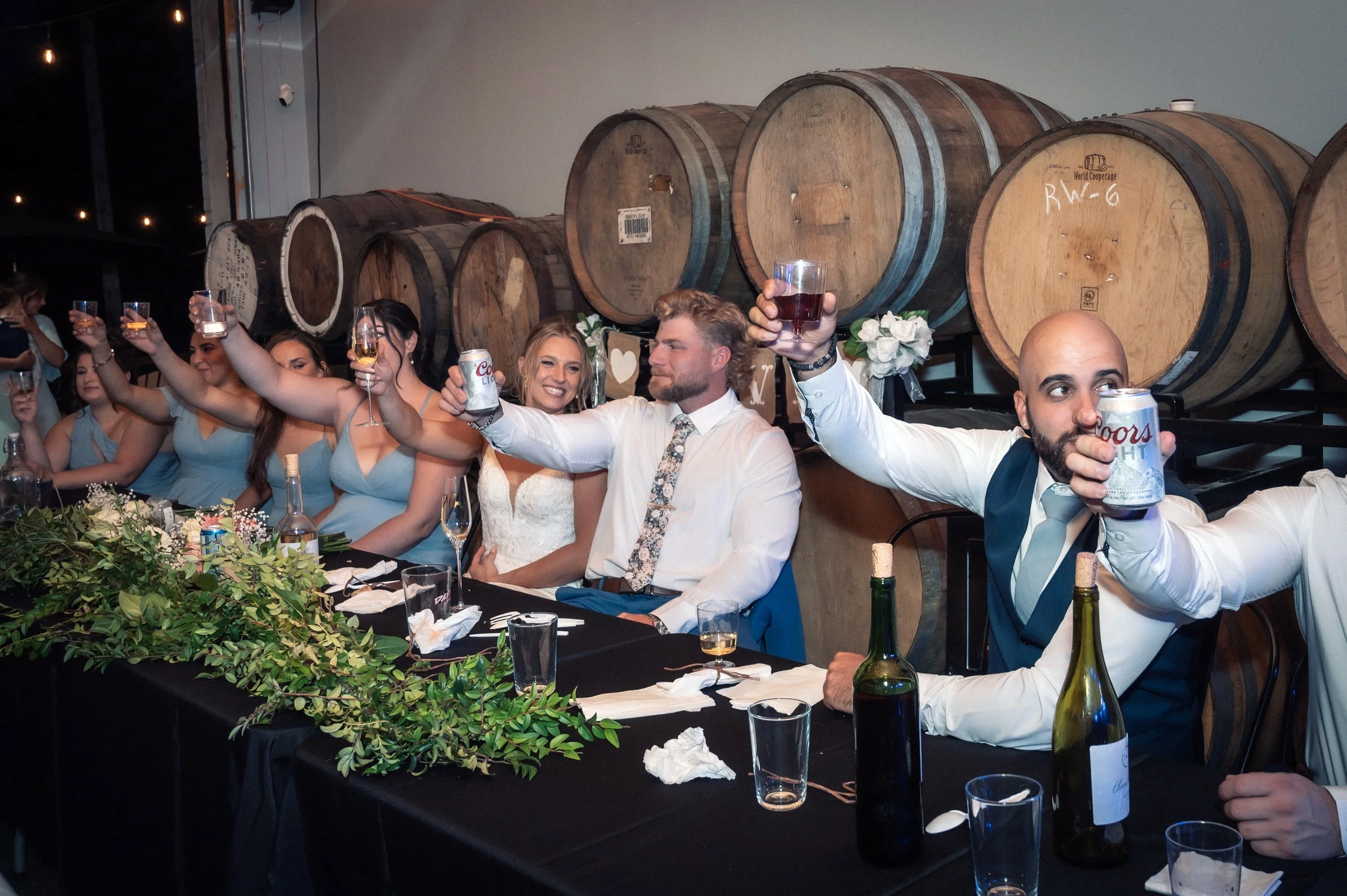 Group of wedding guests sitting at a long table, raising glasses in toast, with wine barrels and floral decorations in the background.