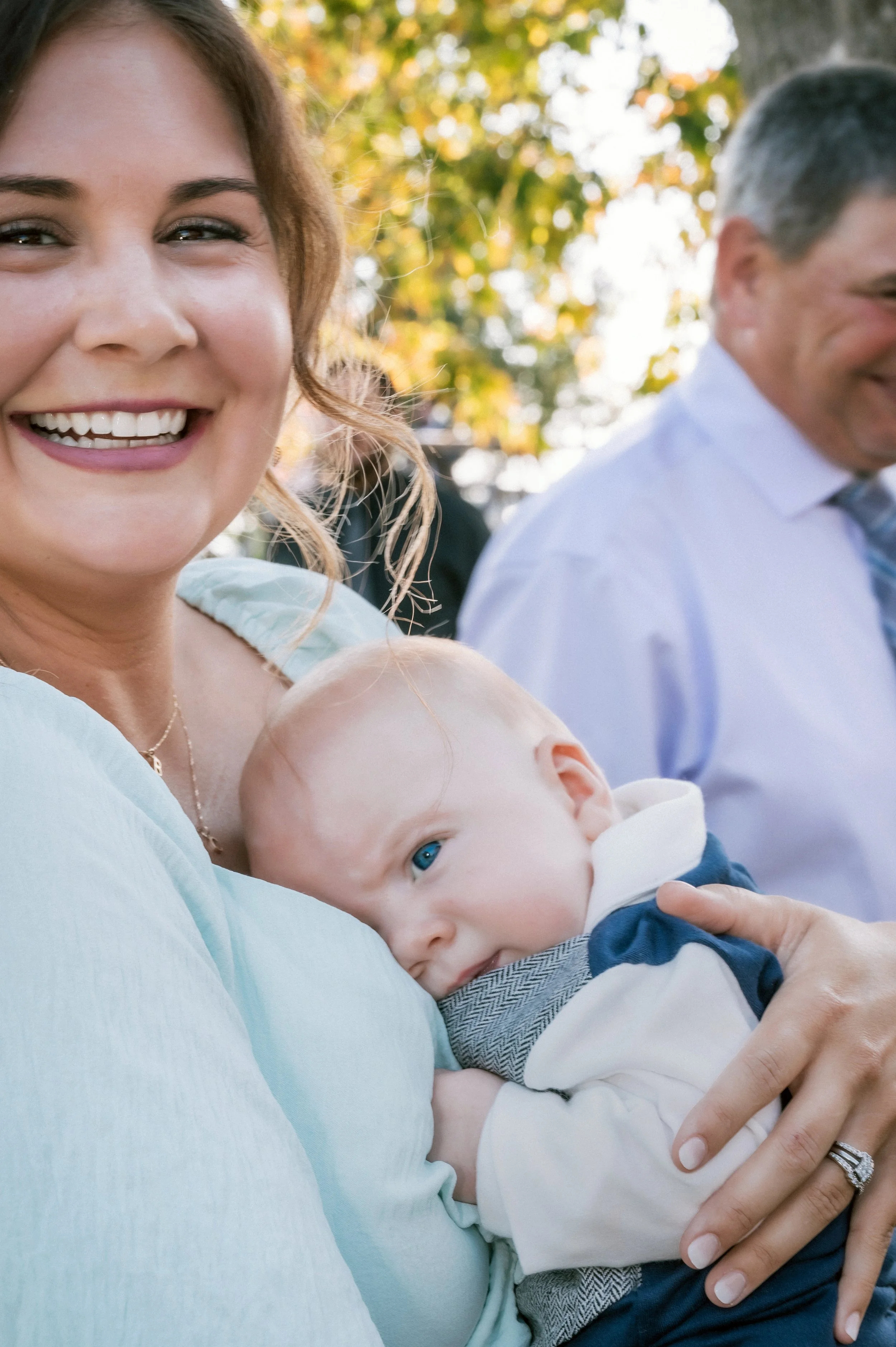 A smiling woman holding a baby with blue eyes, outdoors with autumn foliage in the background.